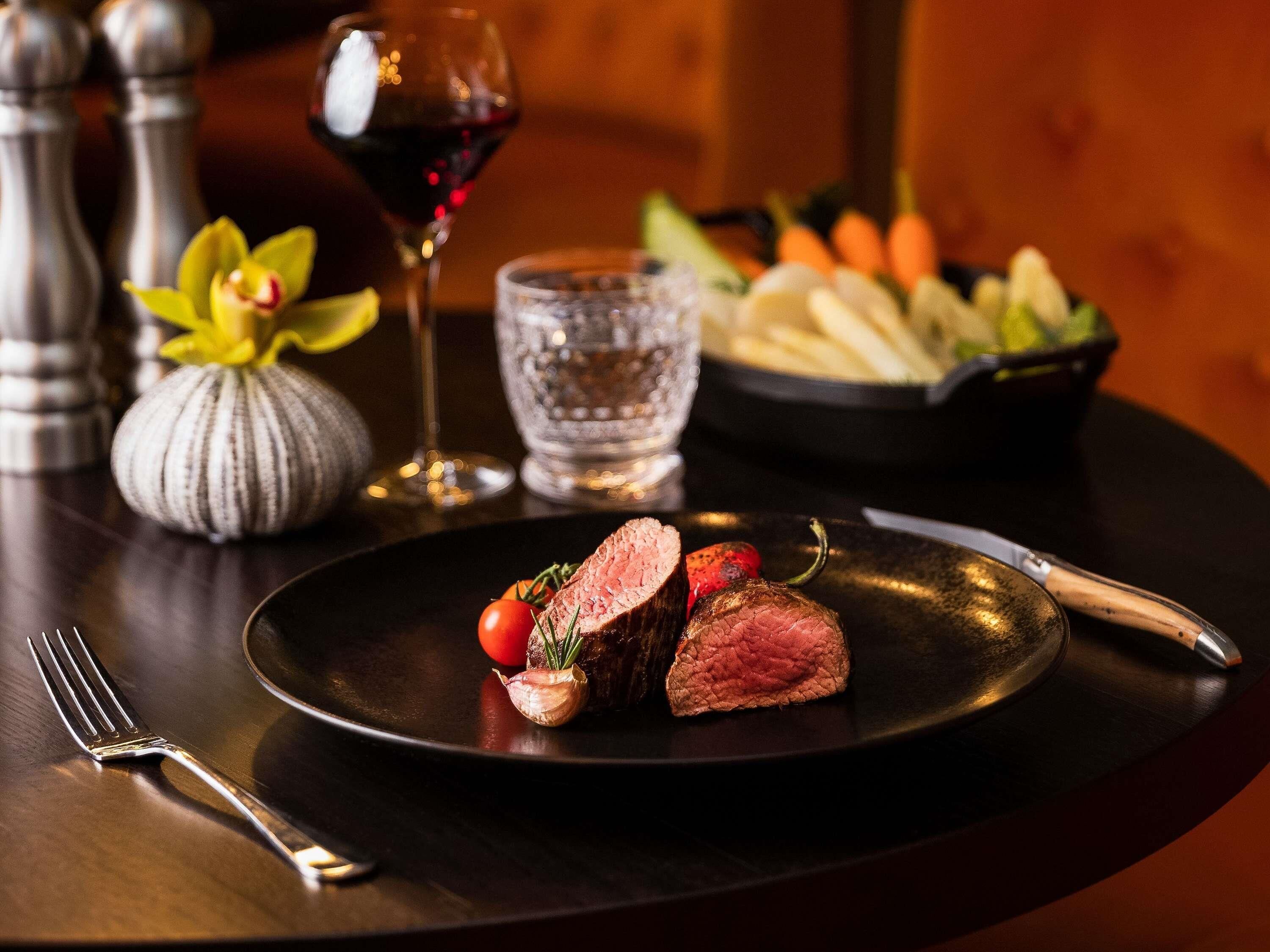 Un plato de carne con verduras, una copa de vino tinto y un bol de vegetales sobre una mesa.