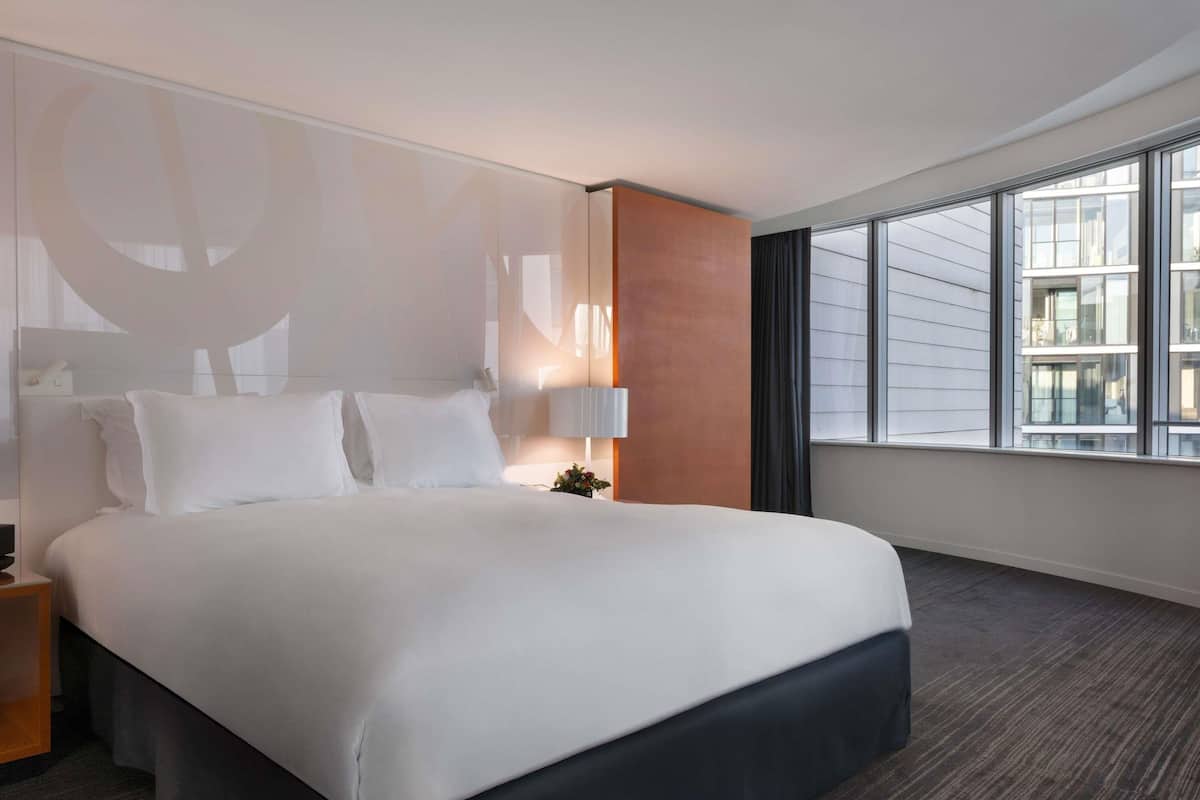 A hotel room with a large bed, a nightstand, a lamp, and a view of the city through large windows.