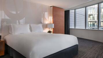 A hotel room with a large bed, a nightstand, a lamp, and a view of the city through large windows.