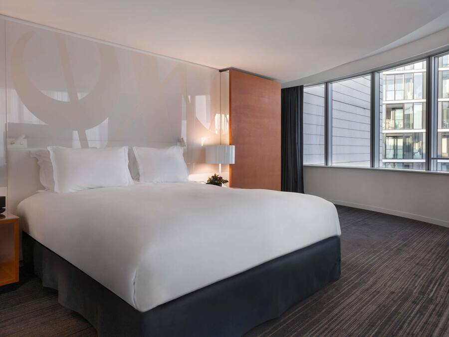 A hotel room with a large bed, a nightstand, a lamp, and a view of the city through large windows.