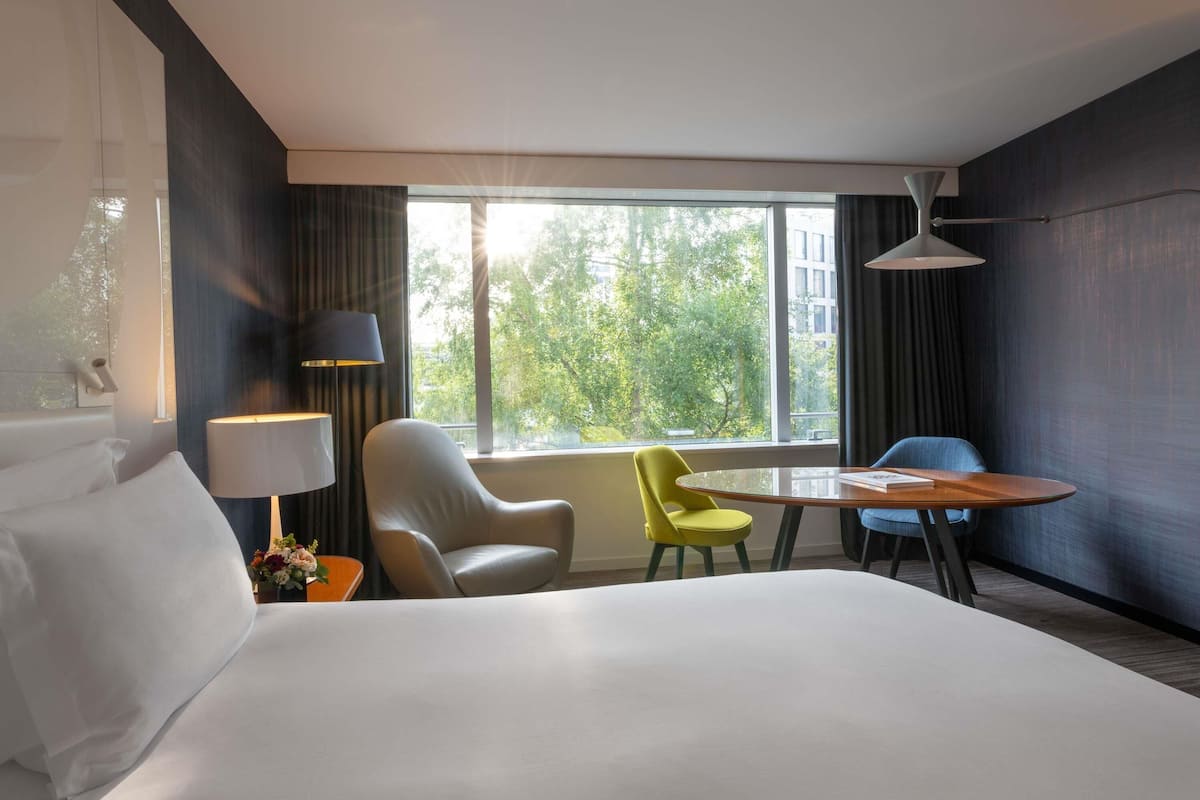Une chambre d’hôtel avec un grand lit, deux chaises, une petite table, une lampe et une fenêtre donnant sur des arbres.