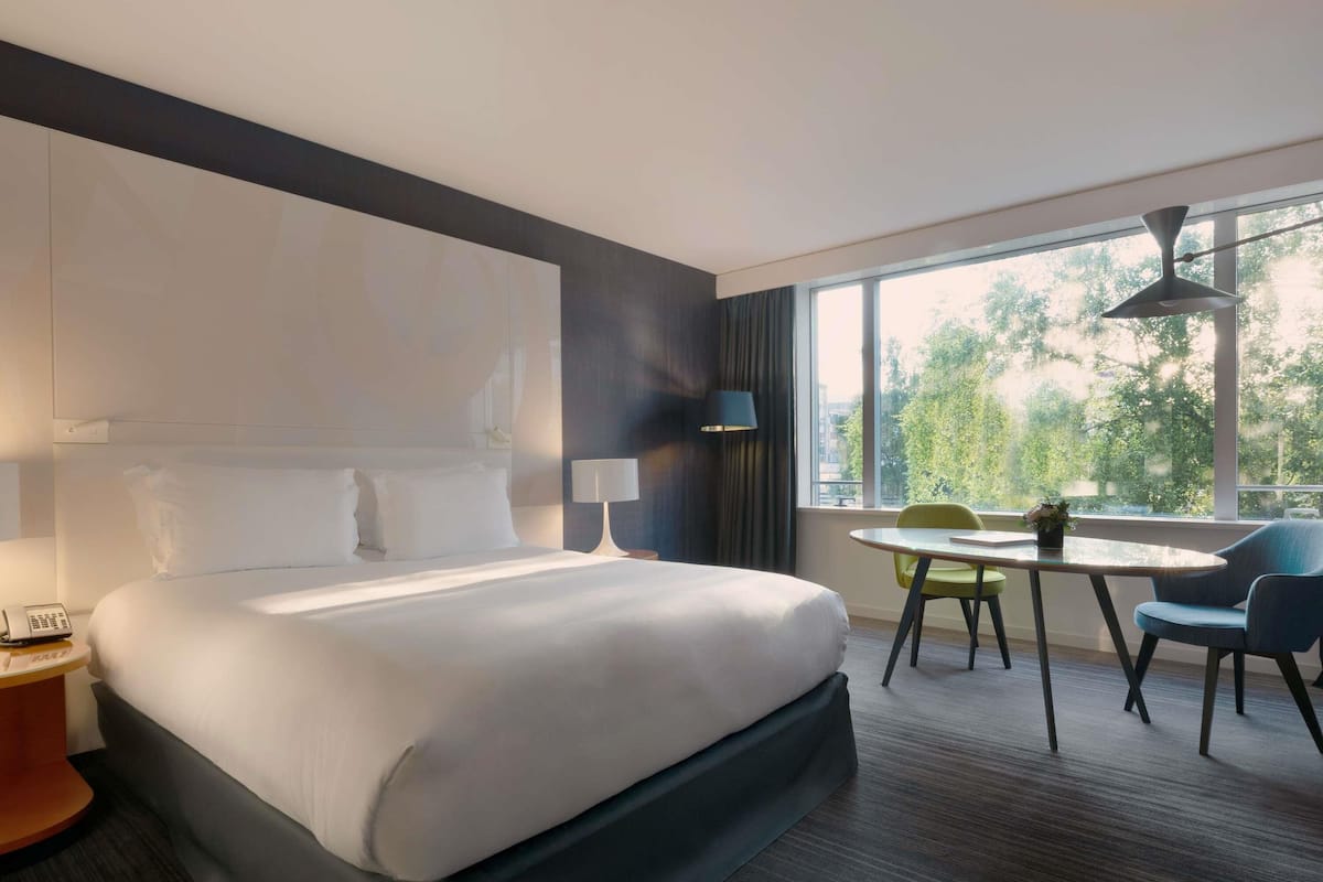 Une chambre d’hôtel avec un grand lit, un bureau, une chaise, une lampe et une vue sur les arbres par la fenêtre.