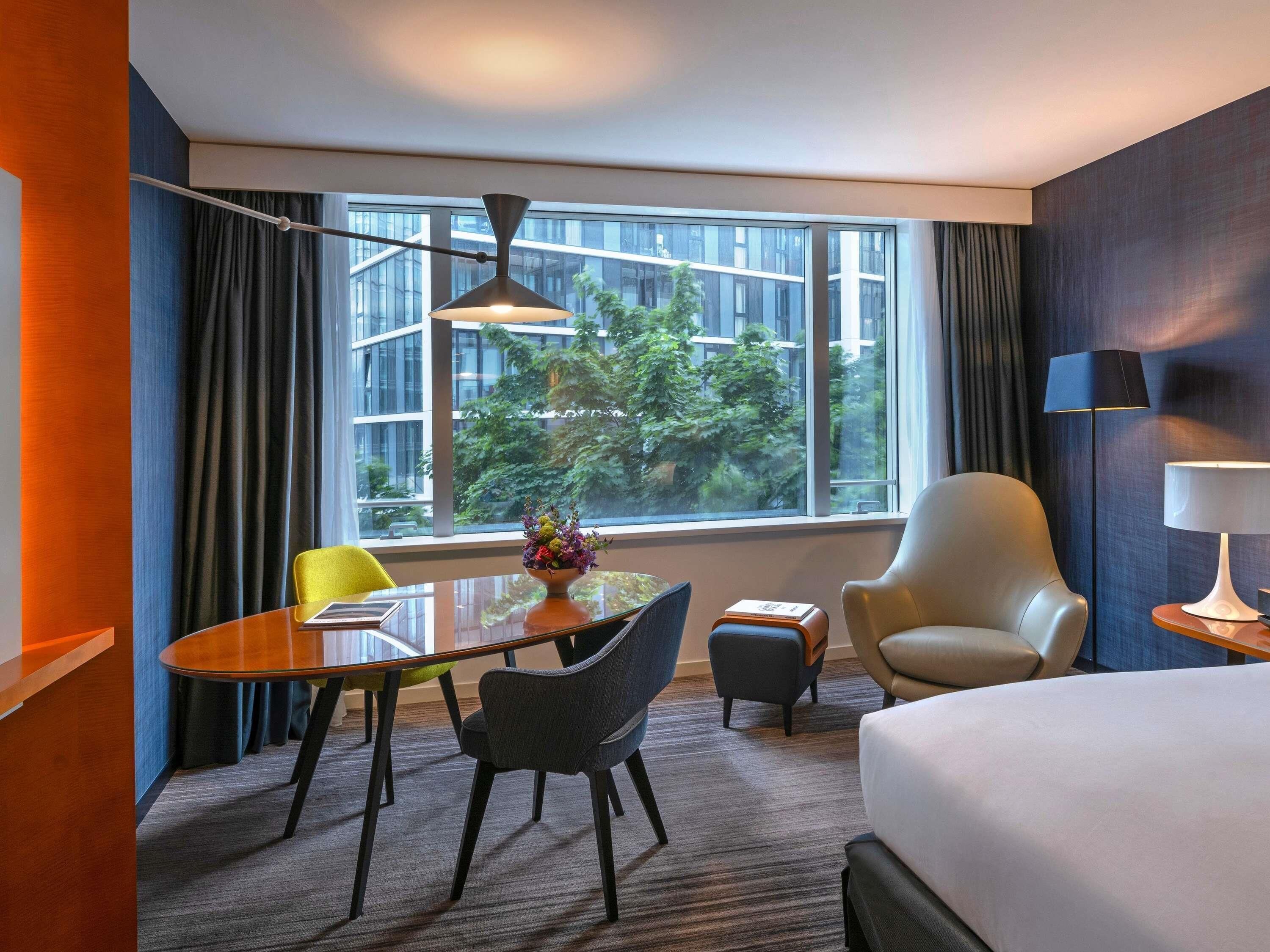 Habitación de hotel con cama, escritorio, silla, mesa, lámpara y vistas a edificios y árboles a través de amplias ventanas.