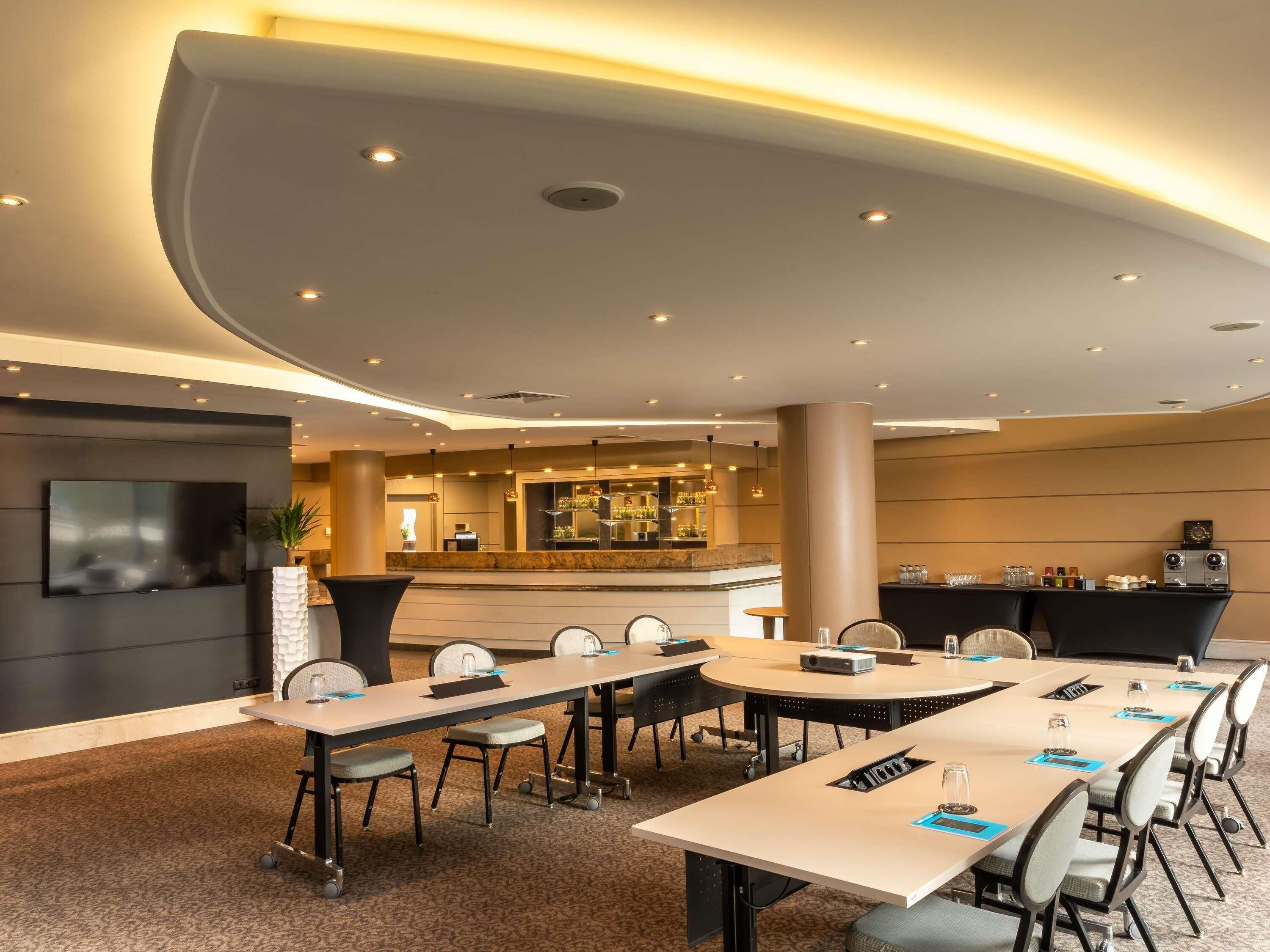 Ein moderner Hotelkonferenzraum mit mehreren Tischen, Stühlen, einer Bar und einem Fernsehbildschirm.
