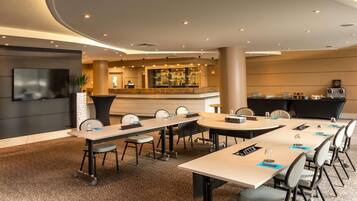 A modern hotel conference room with multiple tables, chairs, a bar area, and a television screen.