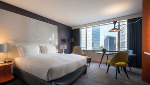 A hotel room with a large bed, bedside tables, a desk with chairs, a window with a city view, and a modern lamp.