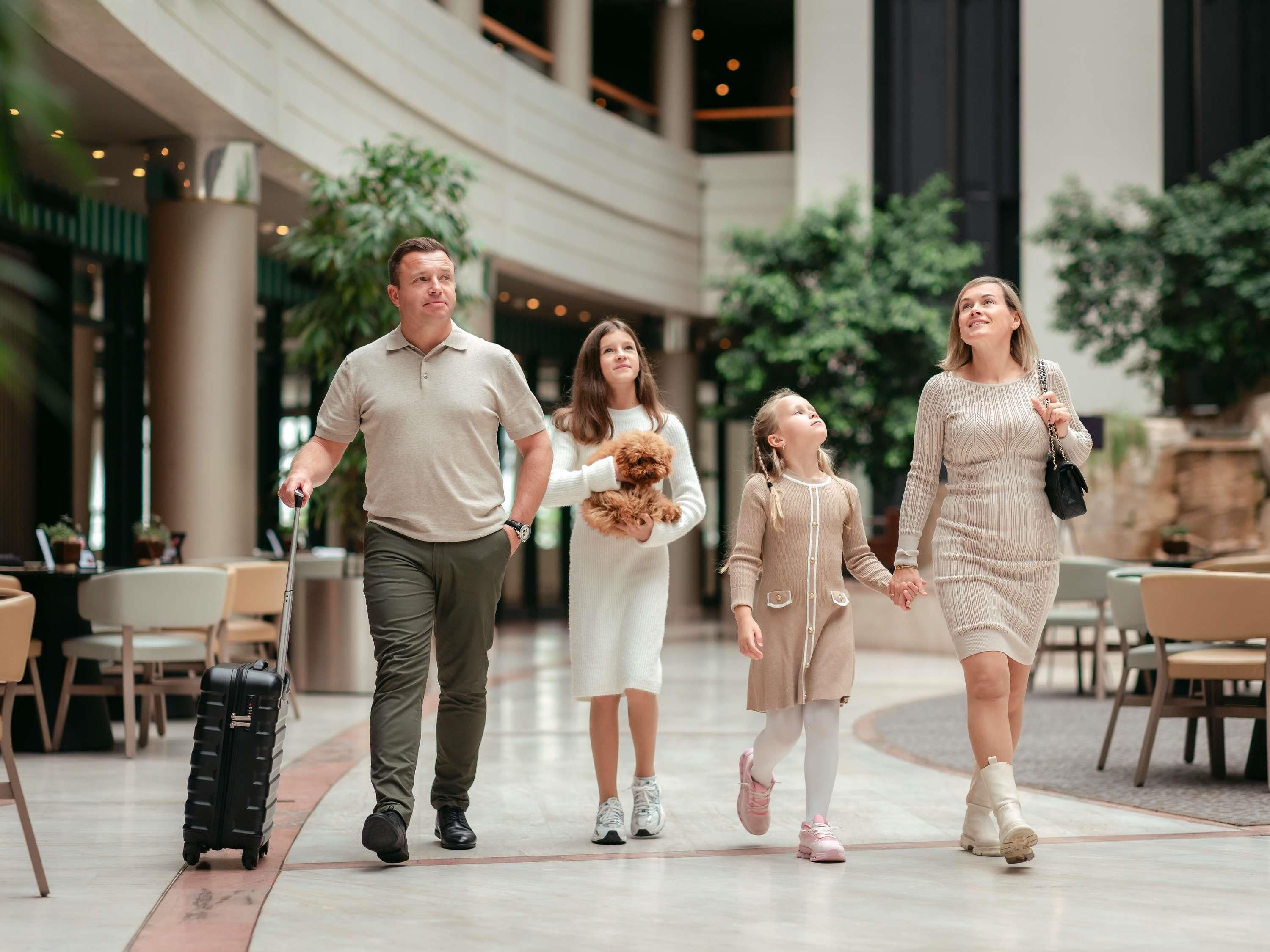 A family walking through a hotel lobby with a dining area and plants in the background.