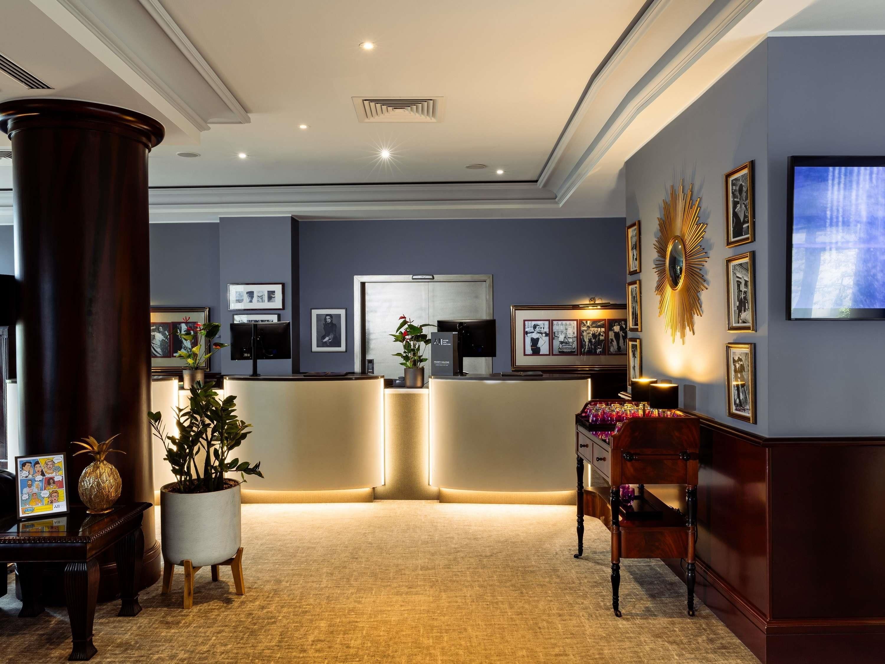 A hotel lobby with a reception desk, framed pictures on the walls, a large potted plant, and a television screen.