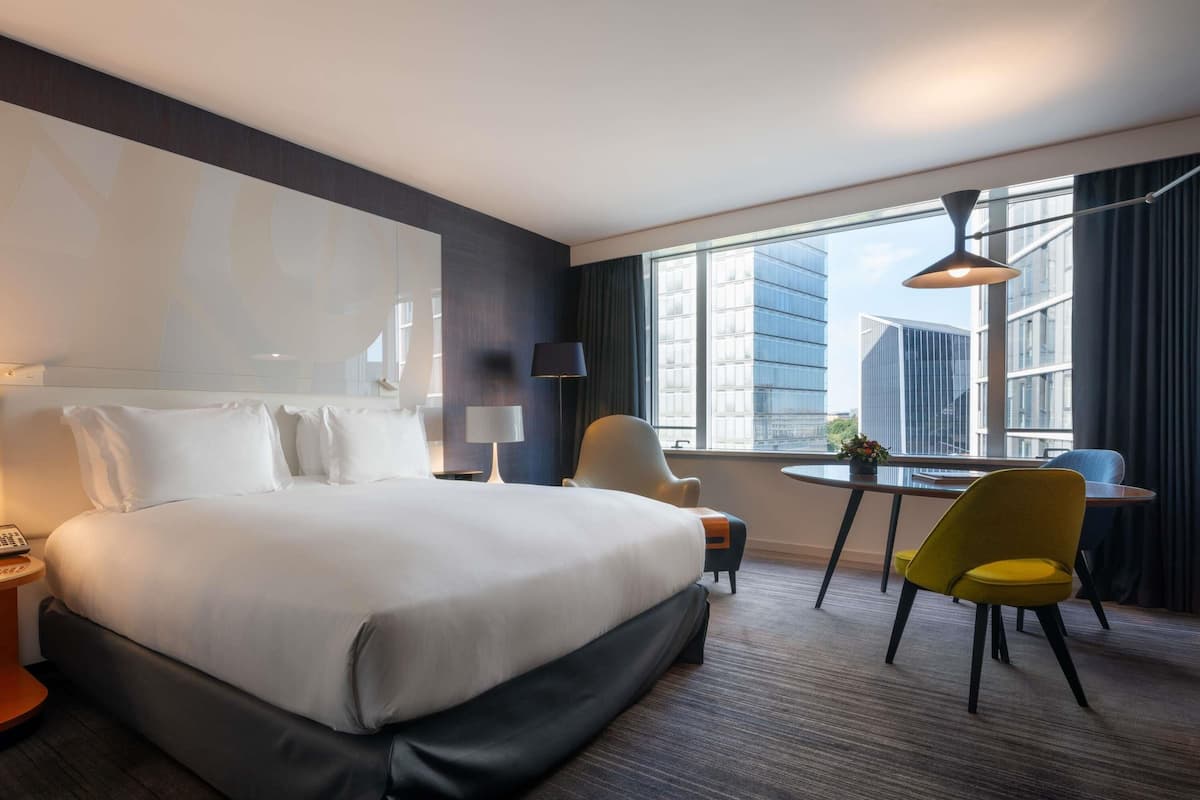 Une chambre d’hôtel avec un grand lit, un bureau, deux chaises, une lampe et une vue sur les immeubles.