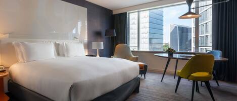 Habitación de hotel con una cama grande, un escritorio, dos sillas, una lámpara y vistas a edificios.