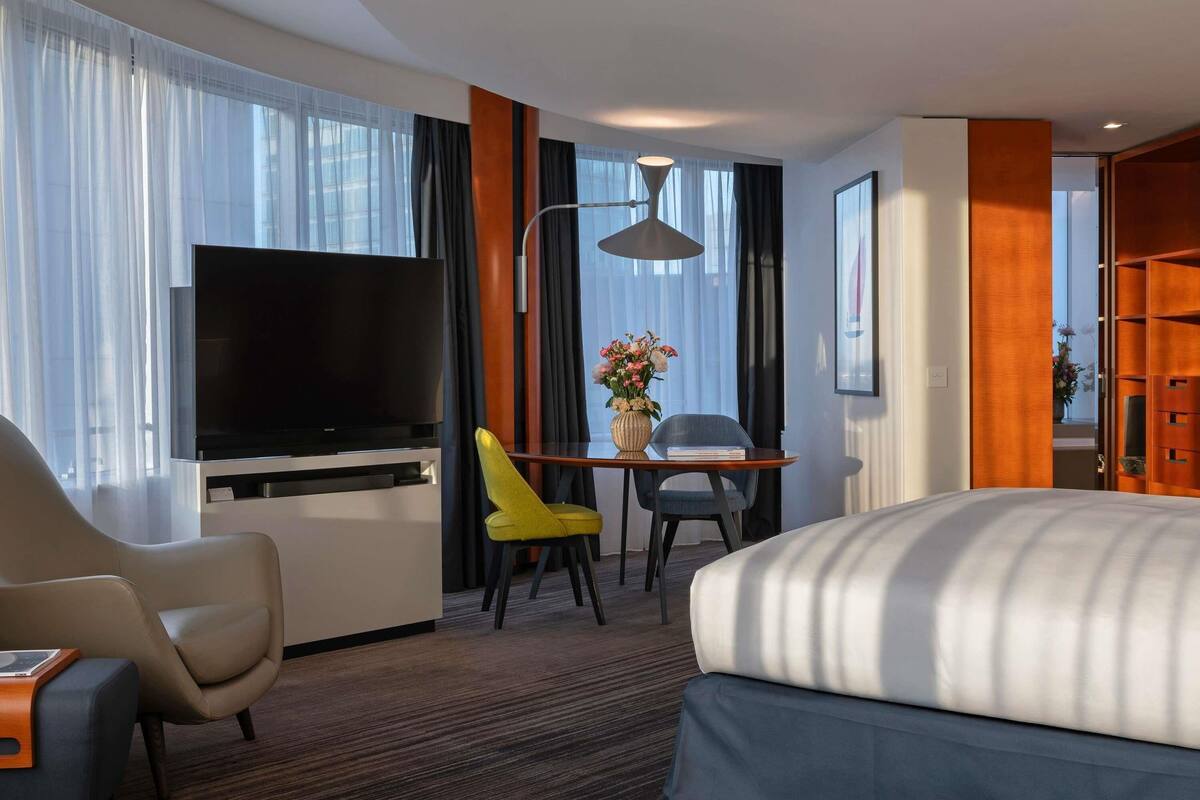 Une chambre d’hôtel comprenant un lit, une télévision, un bureau avec une chaise, une lampe et une fenêtre avec des rideaux.