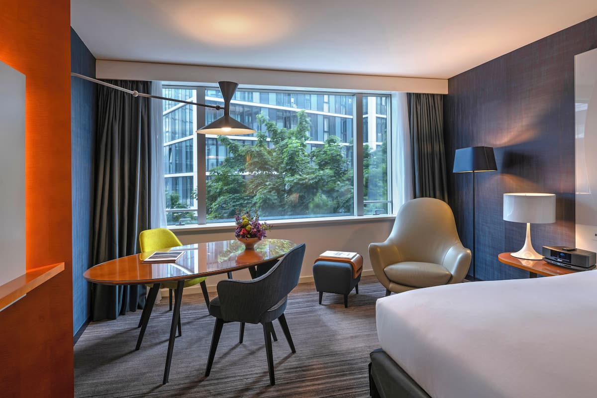 Une chambre d’hôtel avec un lit, un bureau et une chaise, une petite table avec un vase de fleurs, une lampe et une vue sur les arbres par la fenêtre.