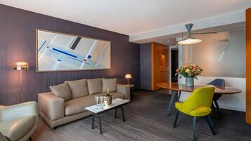 A modern hotel room with a sofa, a dining table with chairs, a coffee table, and a large abstract painting on the wall.