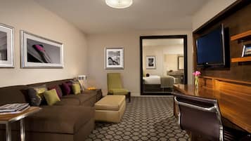 A hotel room with a brown sofa, a green armchair, a wooden desk, a television, and framed pictures on the walls.