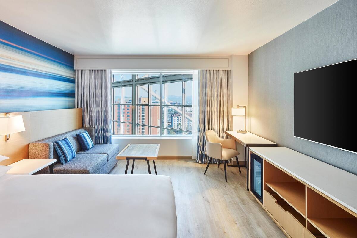 A hotel room with a large bed, a sofa, a desk, a TV, and a window with a city view.