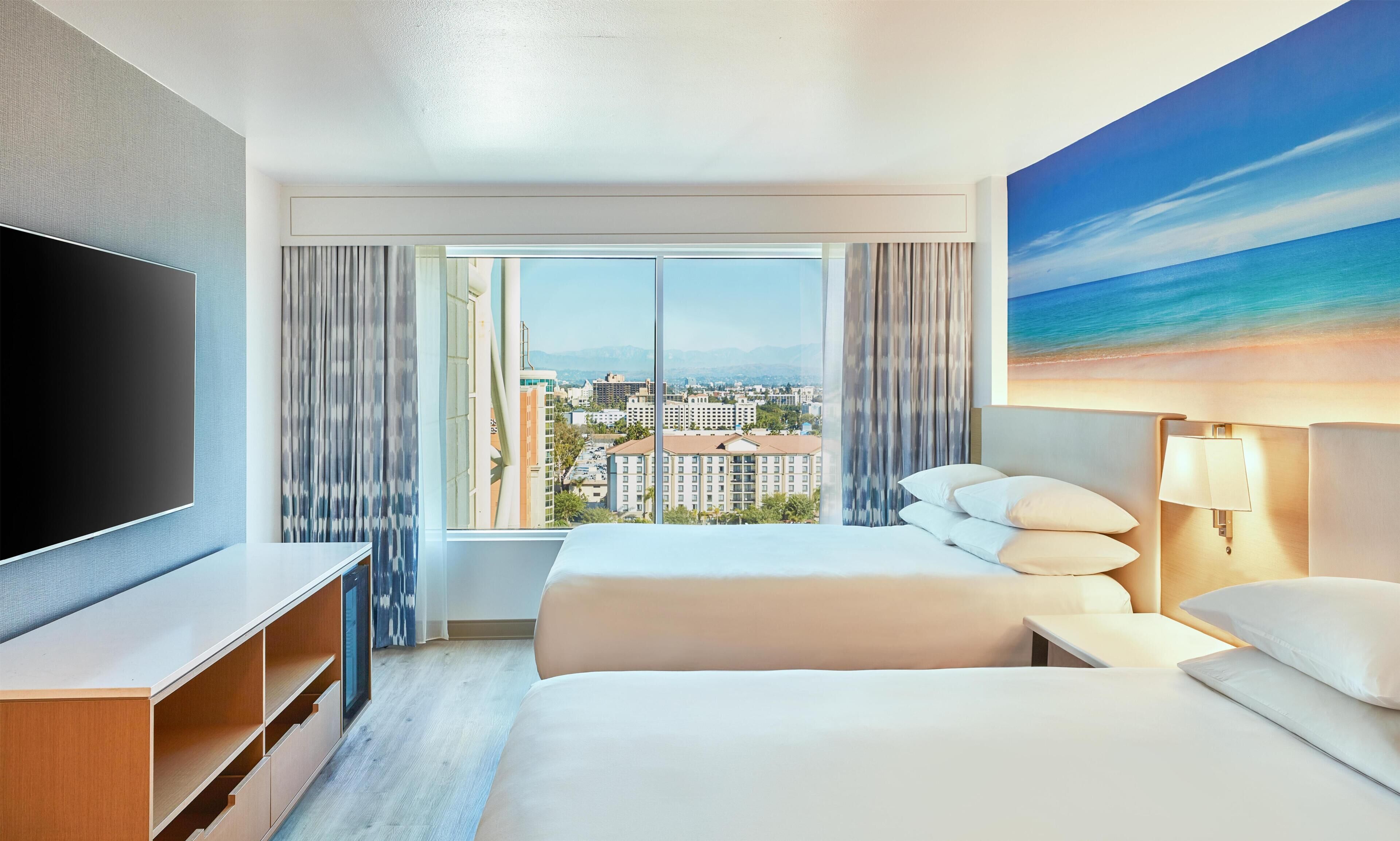 A hotel room with a bed, a TV, a desk, and a large window with a city view.