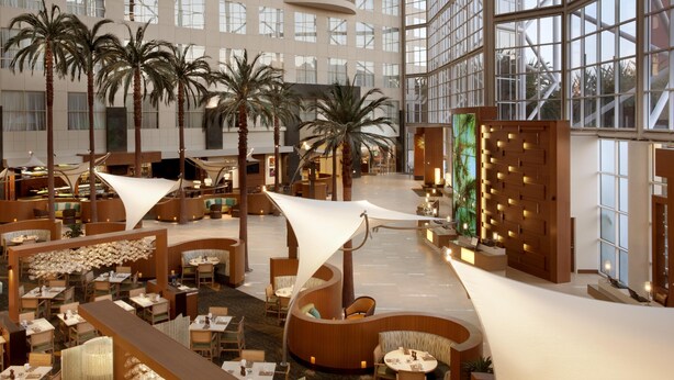 A hotel lobby with multiple seating areas, palm trees, and a large glass facade.