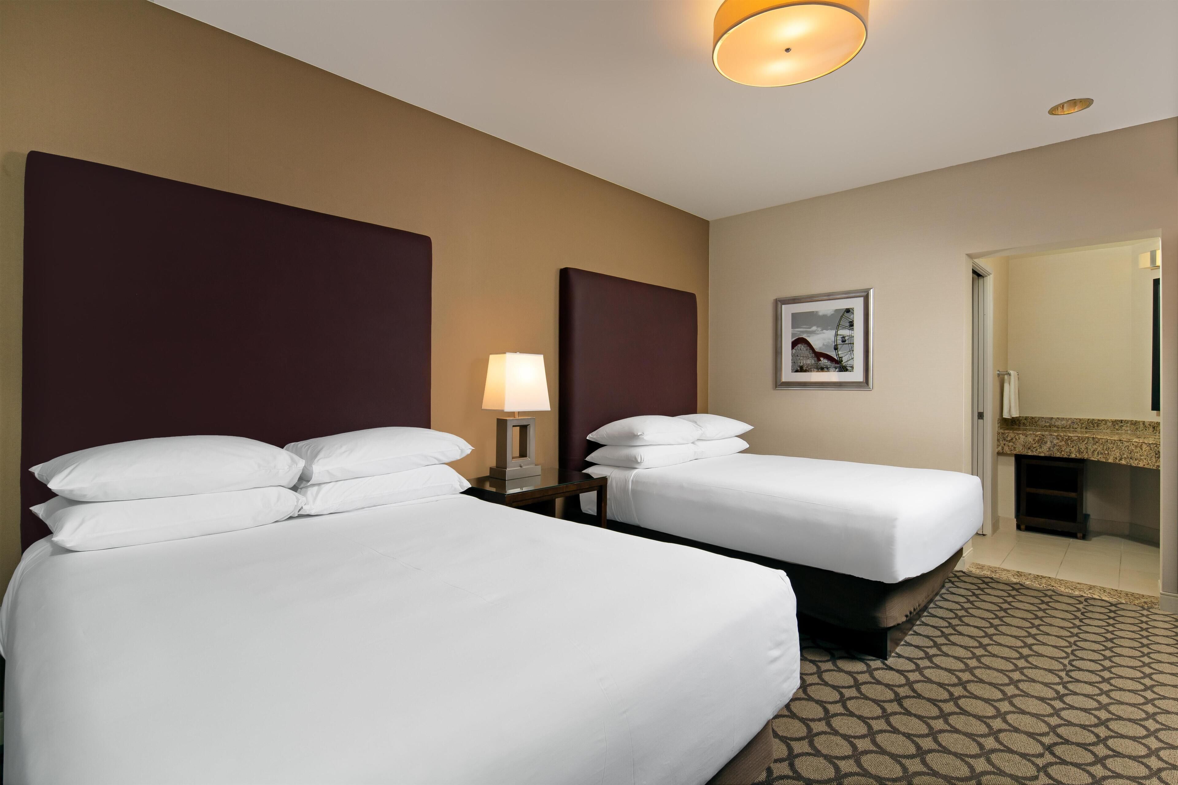 A hotel room with two beds, a nightstand with a lamp, a framed picture on the wall, and a bathroom visible through an open door.