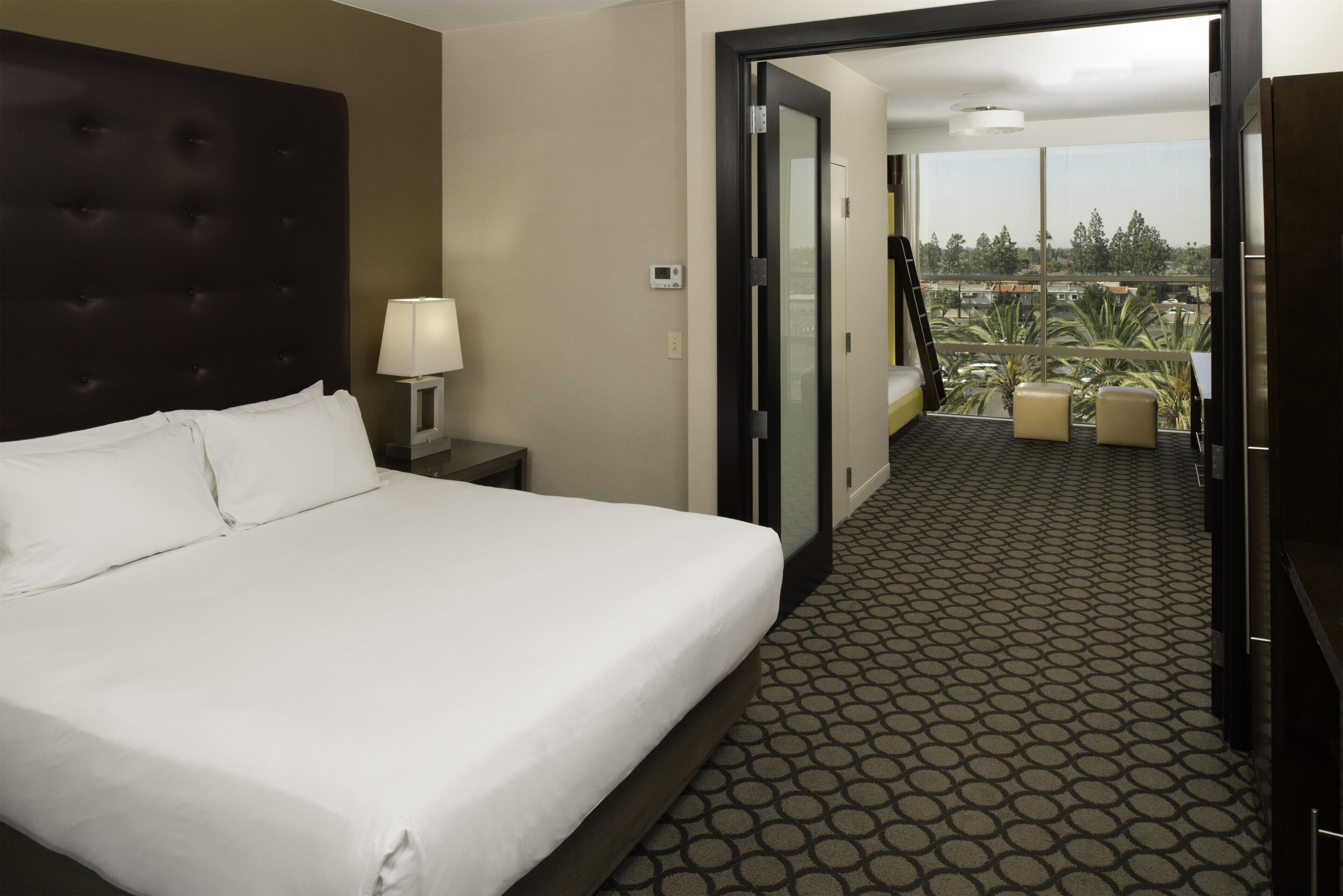 A hotel room with a large bed, a nightstand, a lamp, a digital thermostat, and a view of the outdoors through a glass door.