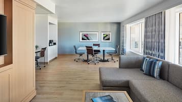 A modern living room with a grey sofa, a coffee table, and a TV on the left. There is a dining area with a white table and chairs, and a kitchen area with cabinets and a plant.