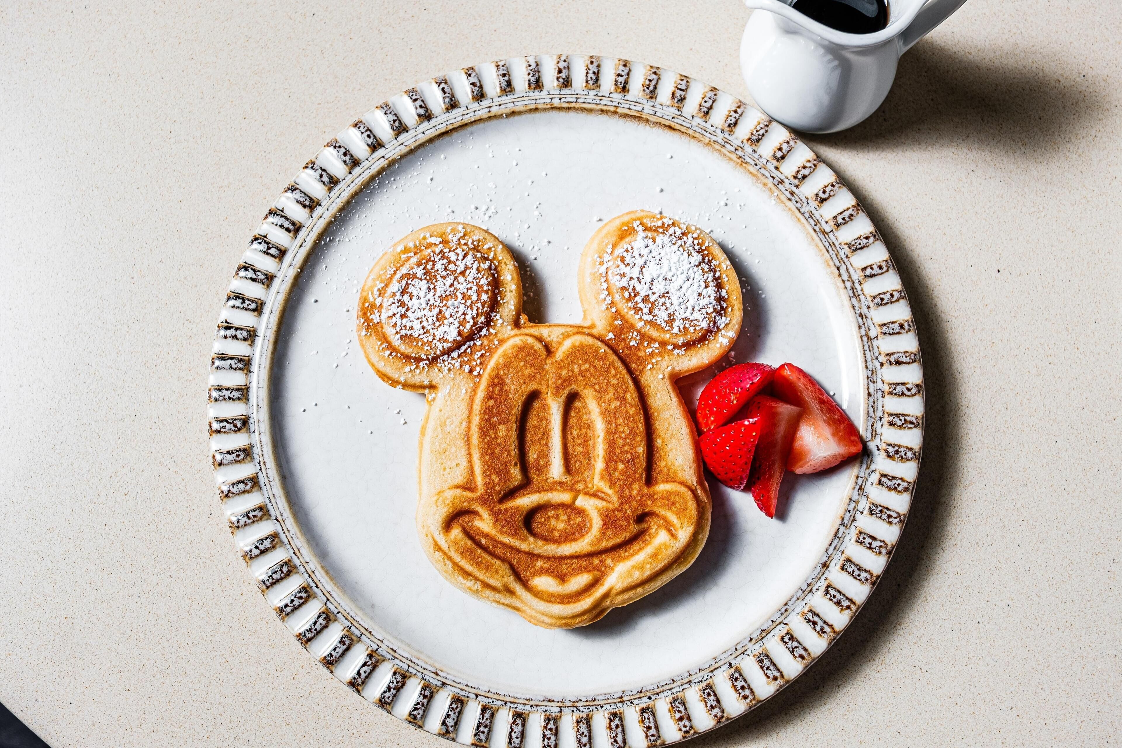 Une assiette en céramique ornée d’un bord décoratif, sur laquelle sont disposées des crêpes en forme de Mickey Mouse et des fraises.