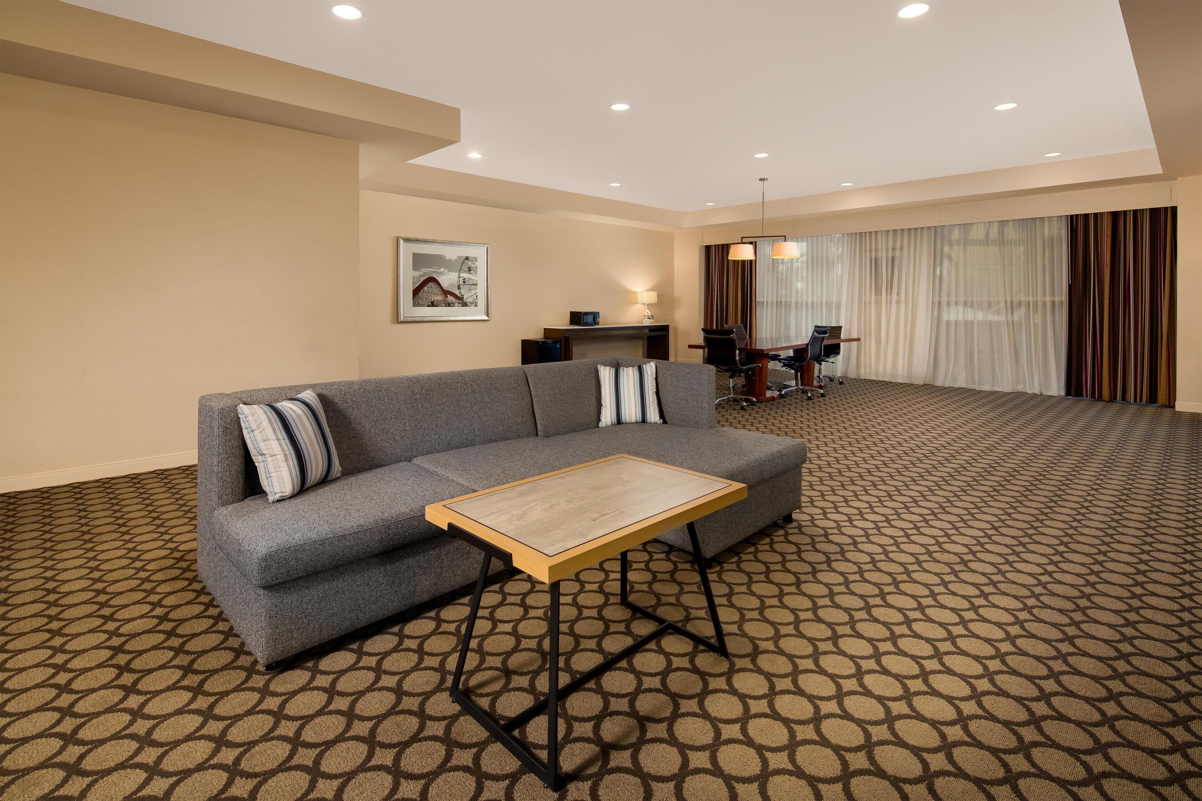 A modern hotel room with a grey sofa, a wooden coffee table, a desk with a lamp, and a dining area with a table and chairs.