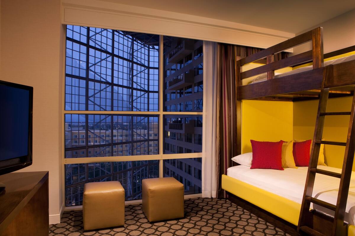 A hotel room with a bunk bed, a television, a large window with a city view, and two ottomans.
