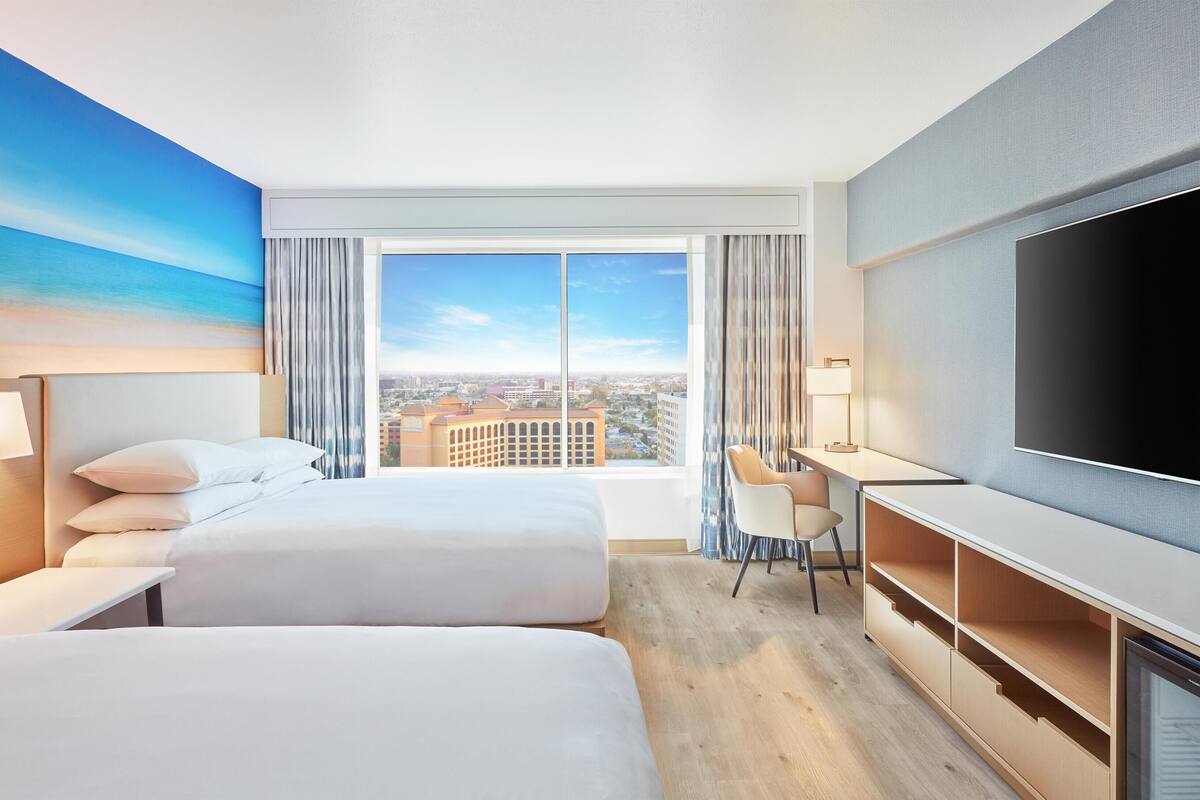 A hotel room with two beds, a desk, a TV, and a large window with a city view.