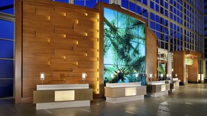 A modern hotel lobby with wooden reception desks, large windows, and a digital screen displaying a tropical scene.