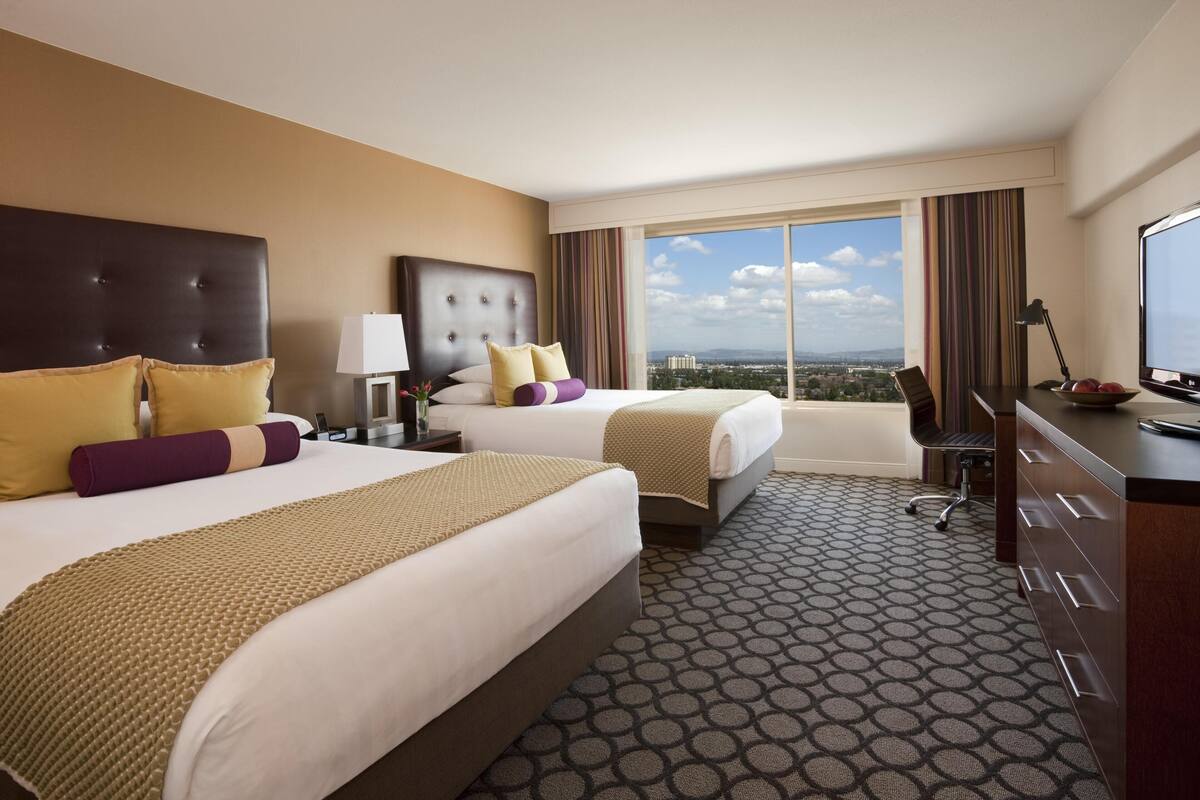 A hotel room with two beds, a desk, a chair, a TV, and a large window with a city view.
