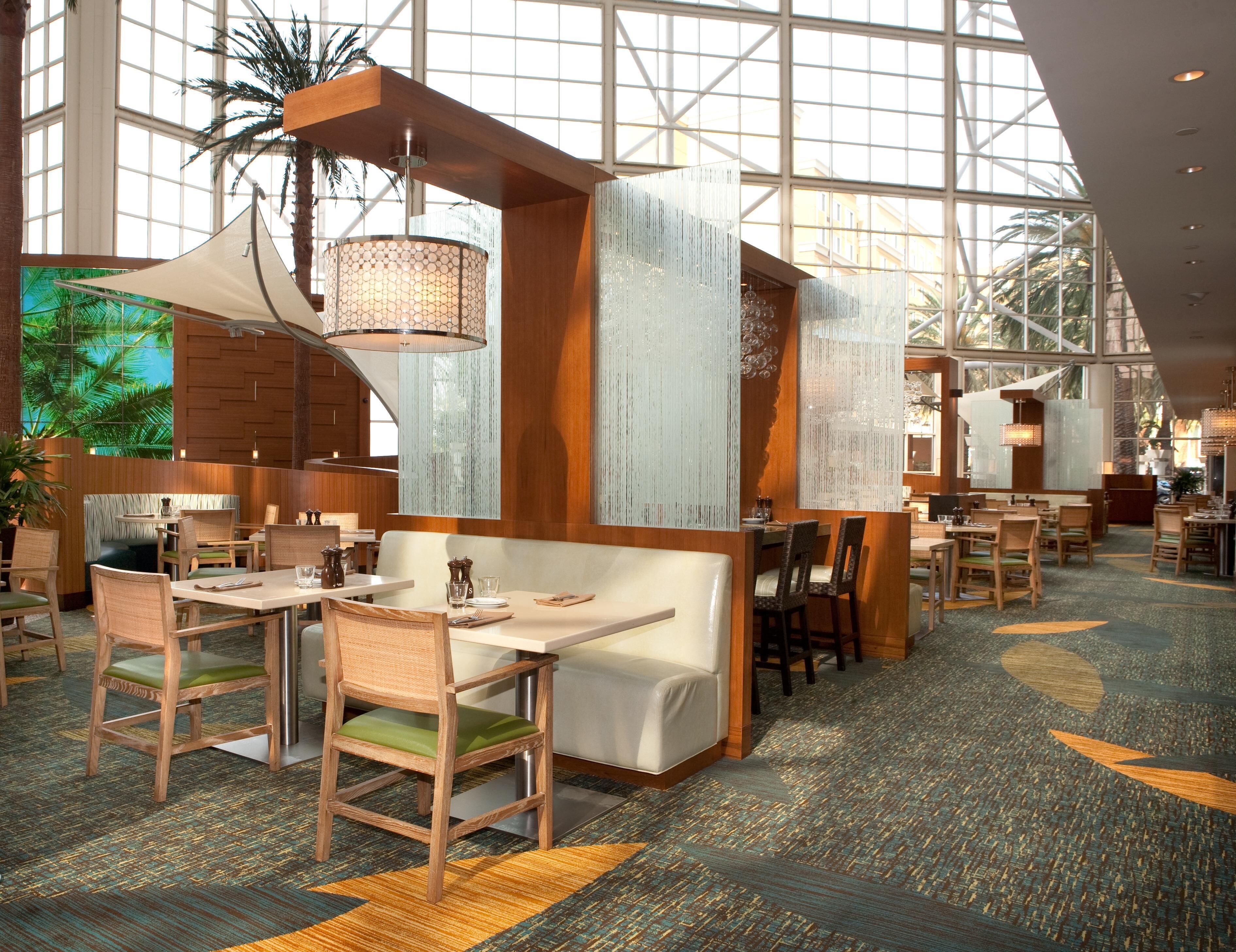 A spacious hotel dining area with high ceilings, large windows, and a tropical view.