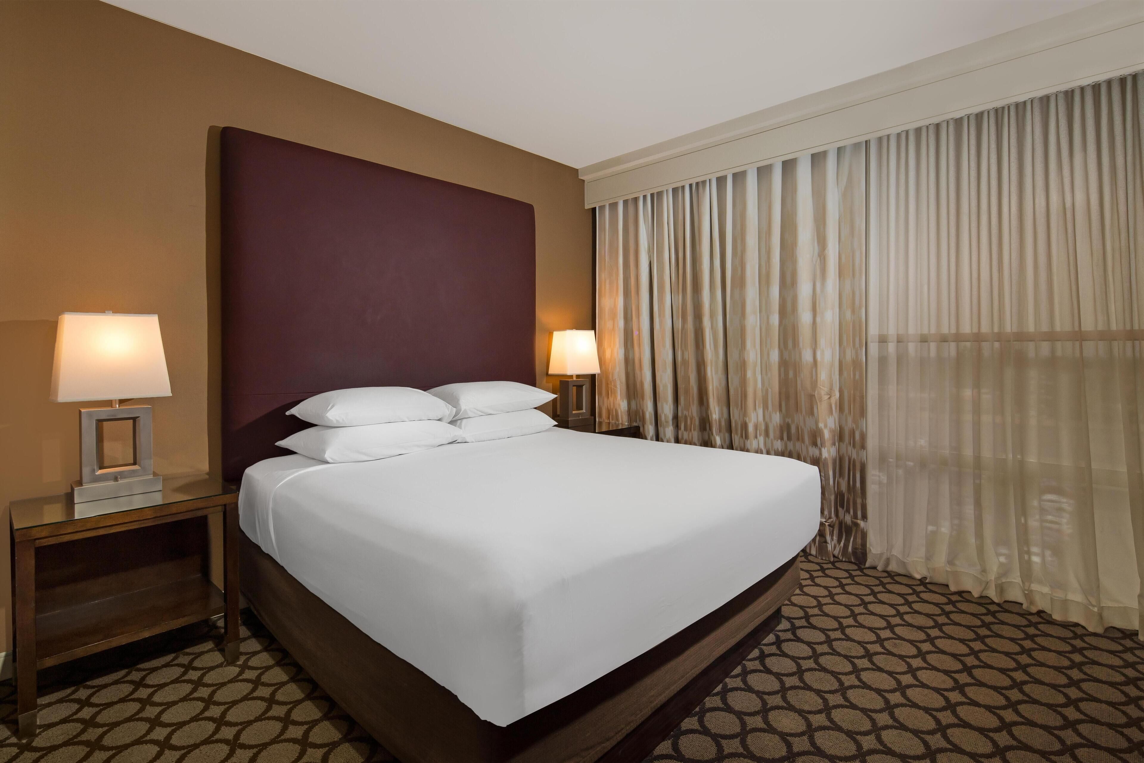 A hotel room with a large bed, two bedside tables with lamps, a headboard, and a window with sheer curtains.