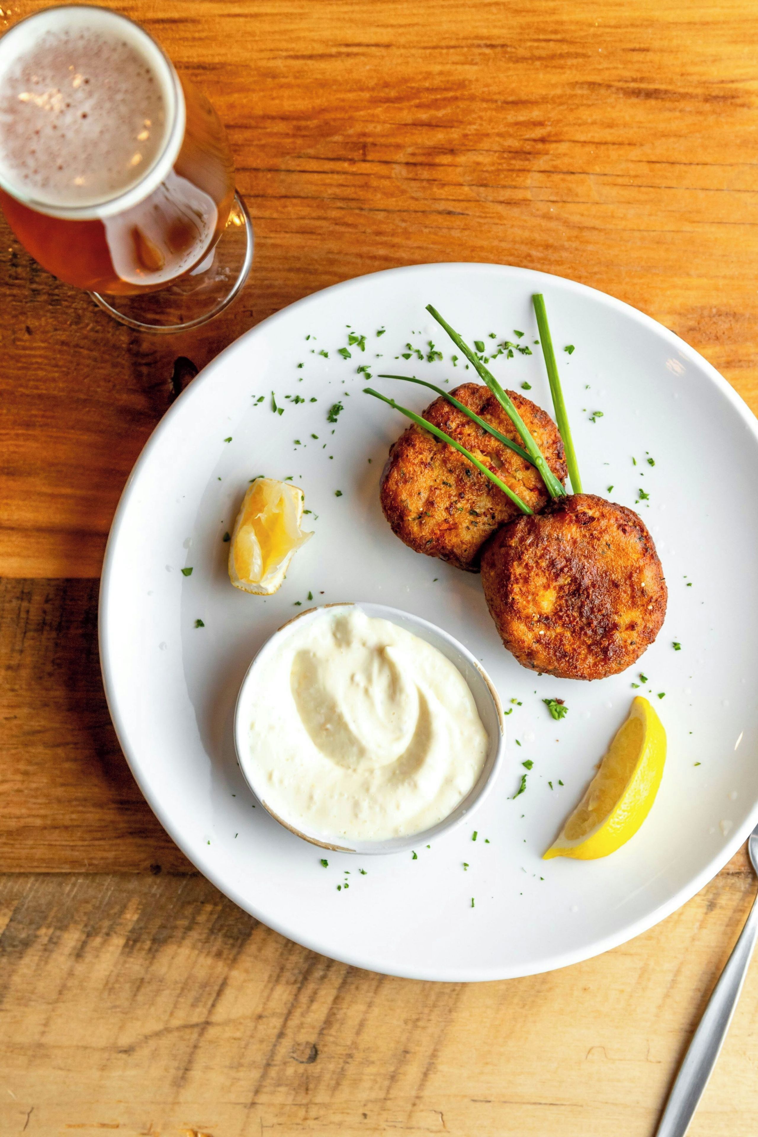 A plate with two golden-brown patties, a dollop of creamy sauce, a lemon wedge, and a glass of beer on a wooden table.