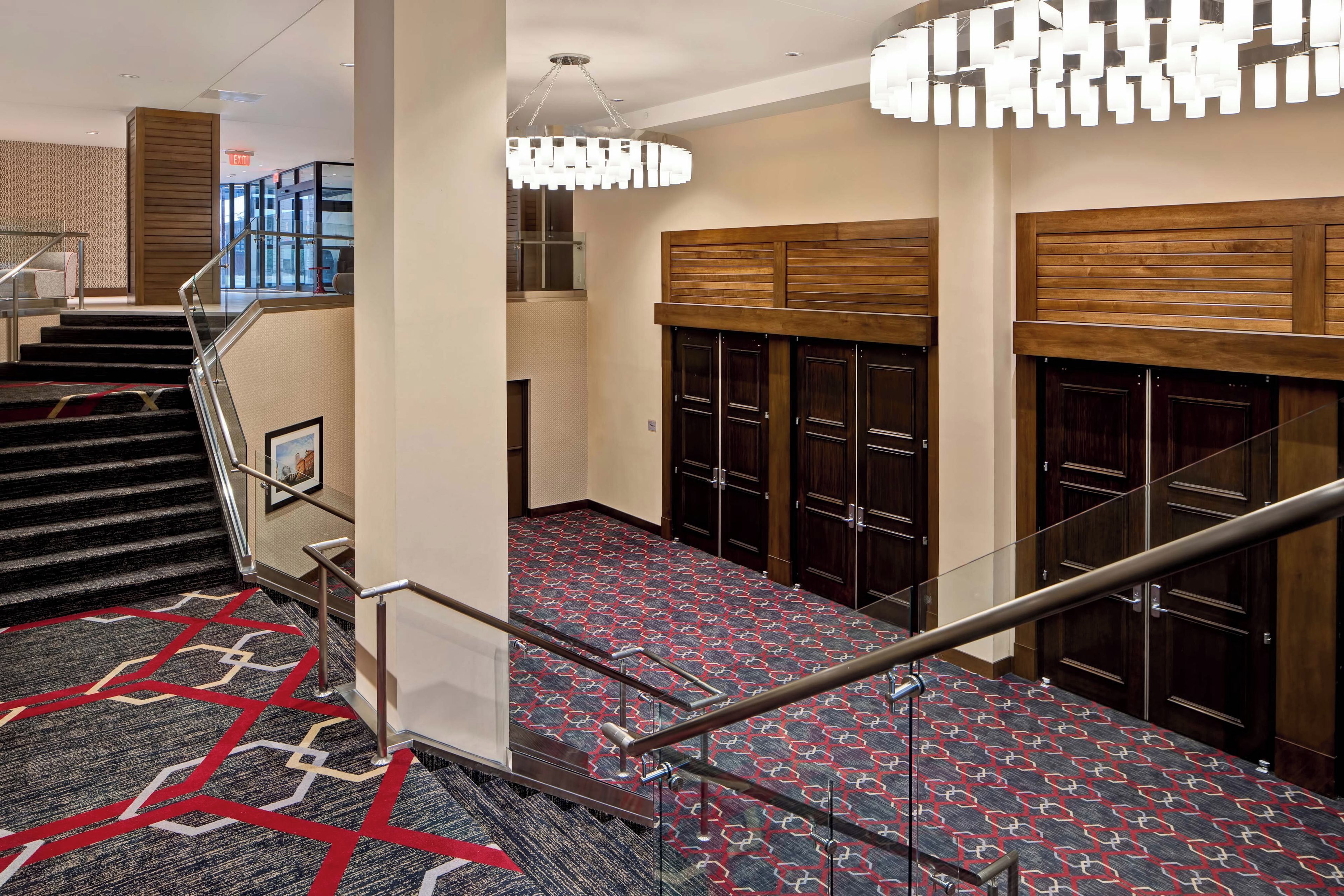 Een hotelhal met een trap, een getextureerd tapijt, houten kasten en een moderne kroonluchter.