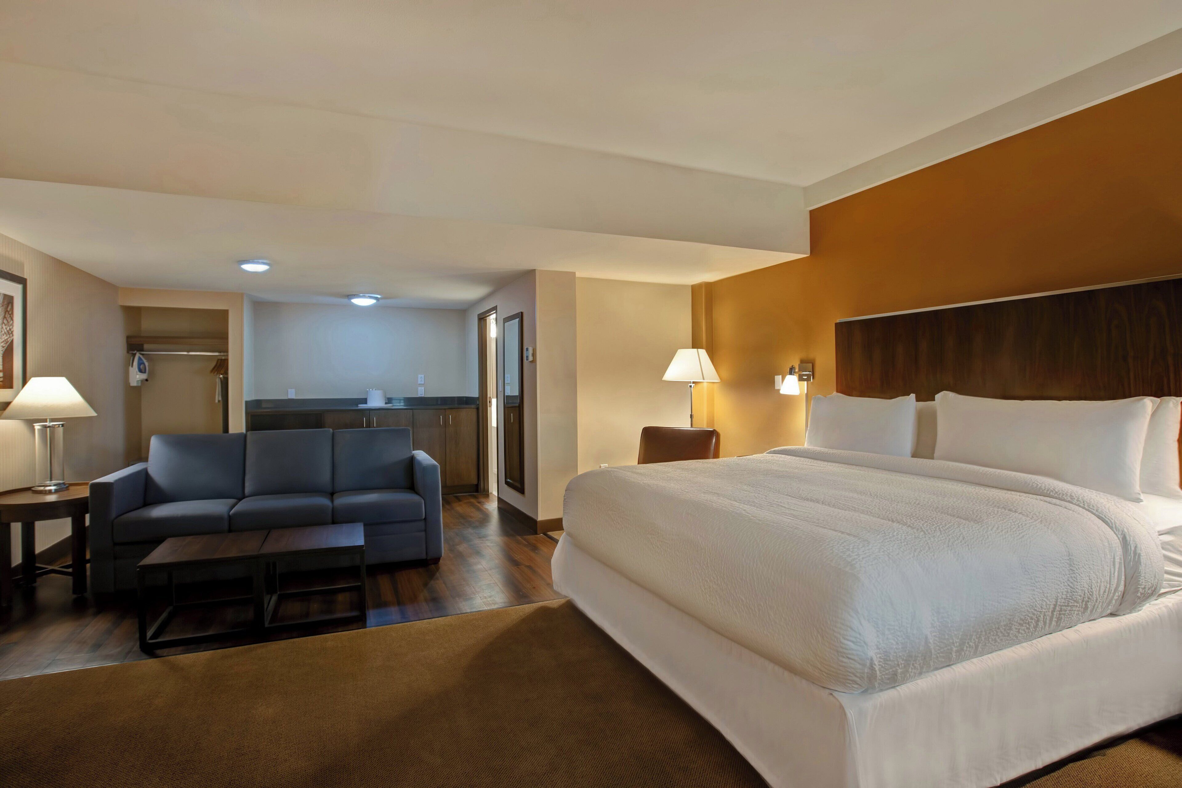 A hotel room with a large bed, a sofa, a coffee table, a lamp, and a view of the kitchen and bathroom.