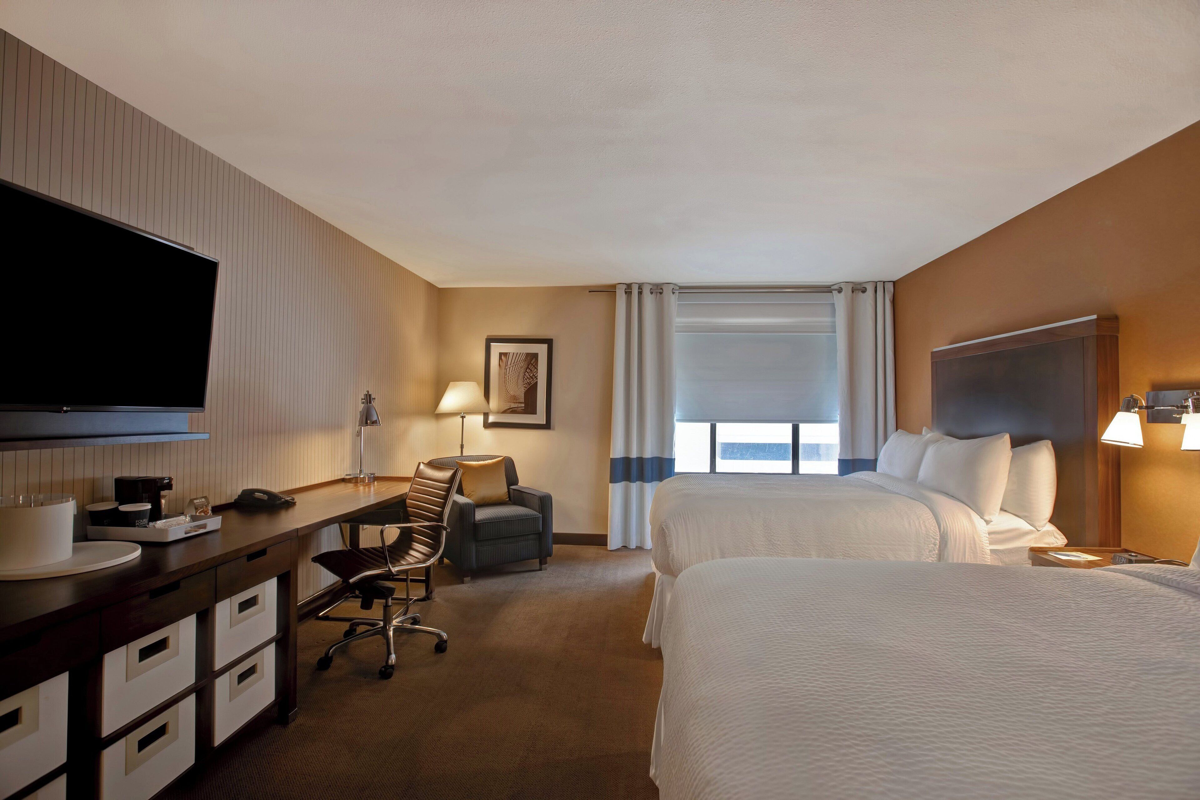 Una habitación de hotel con escritorio, dos camas, televisor y ventana con cortinas.