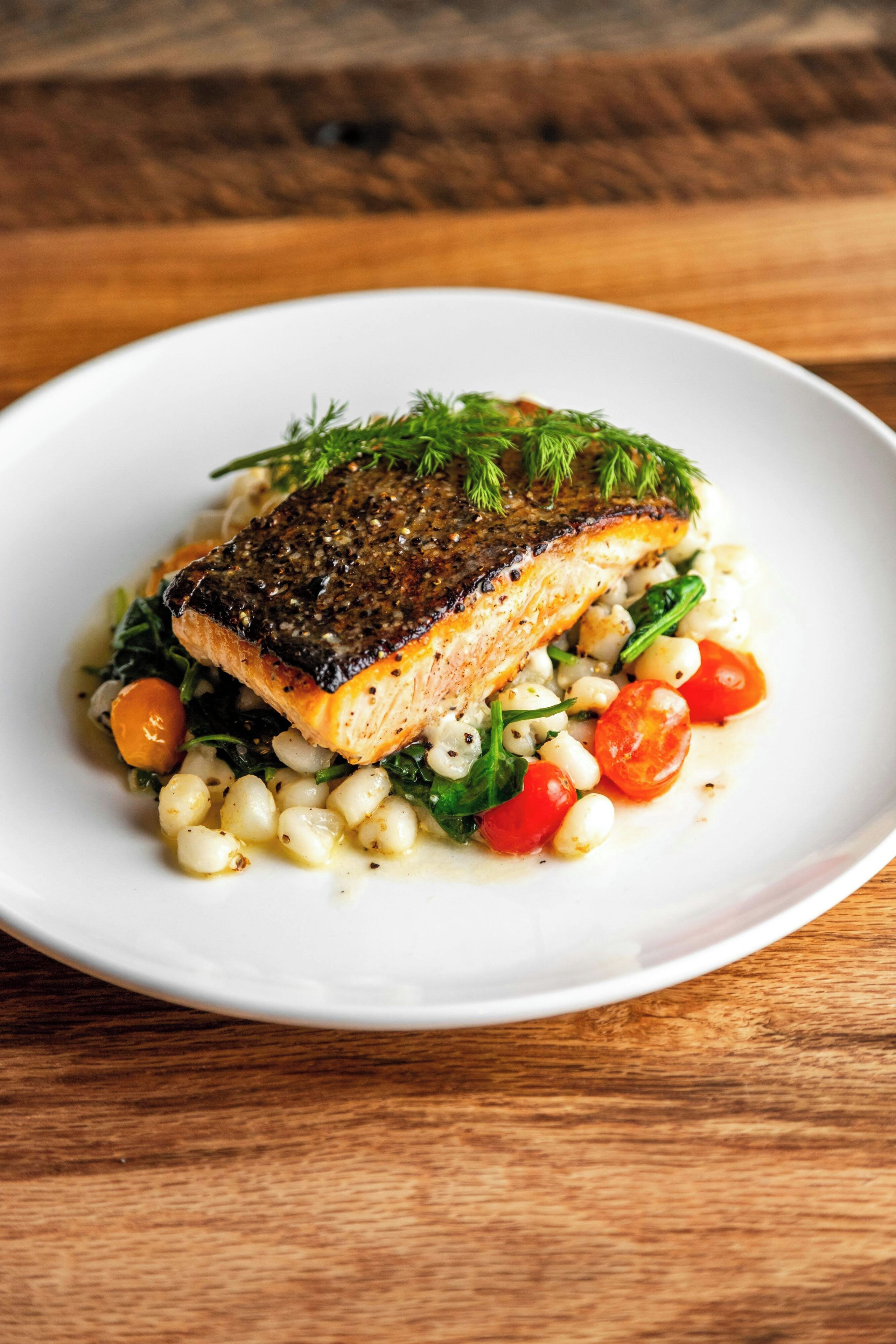 A white plate with a piece of salmon, garnished with dill, served with vegetables and a sauce.