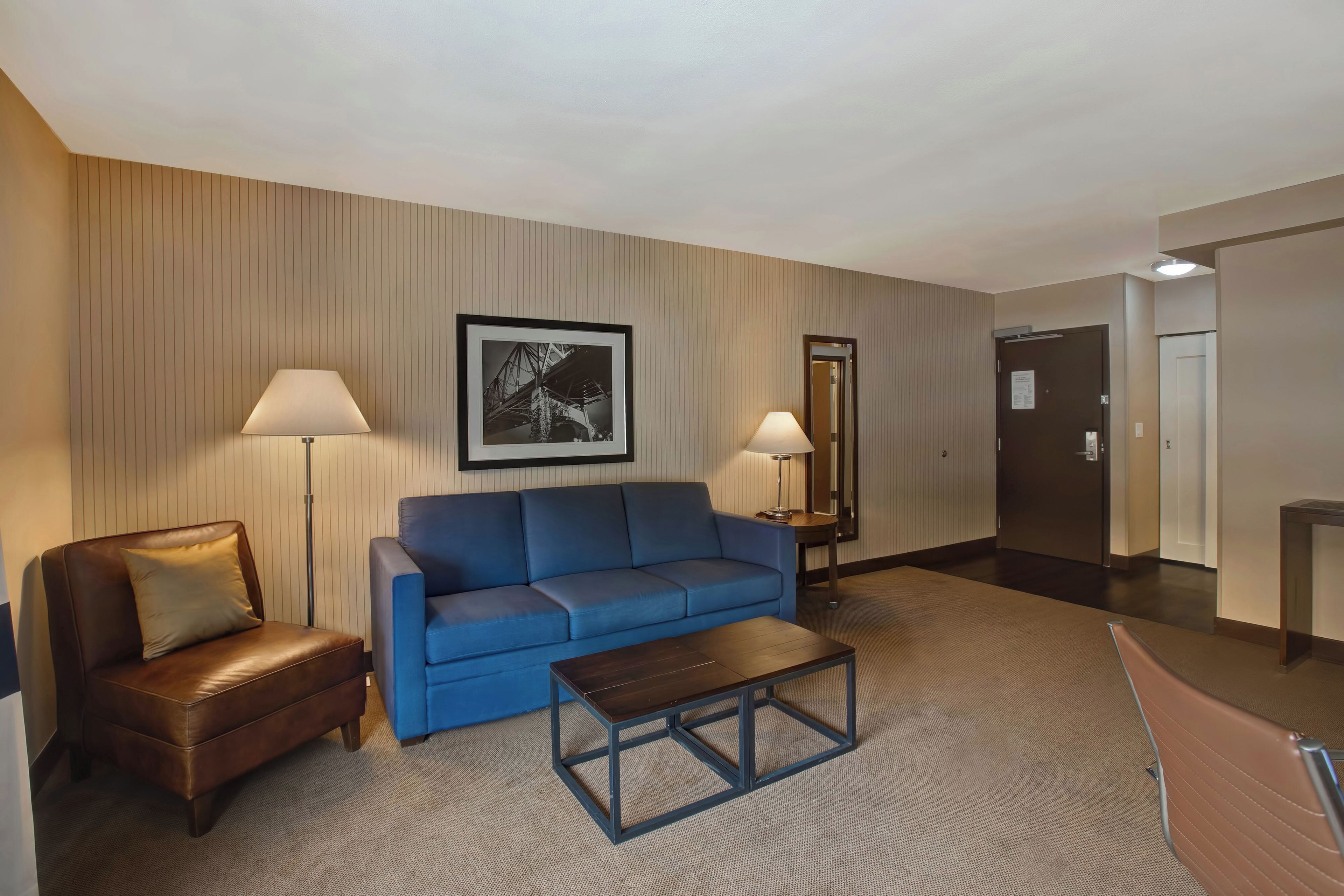 A hotel room with a brown leather chair, a blue sofa, a small wooden coffee table, and two floor lamps.