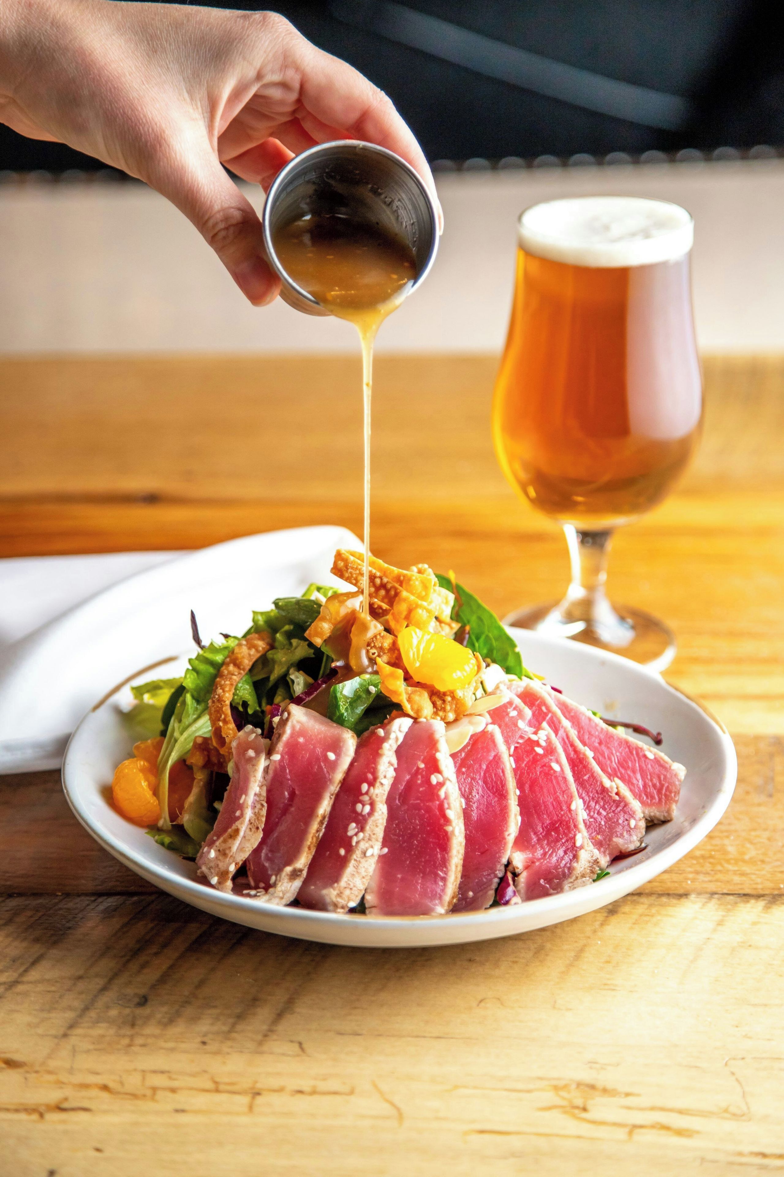 Un plato de comida con una mano sirviendo salsa, una jarra de cerveza y una mesa de madera.