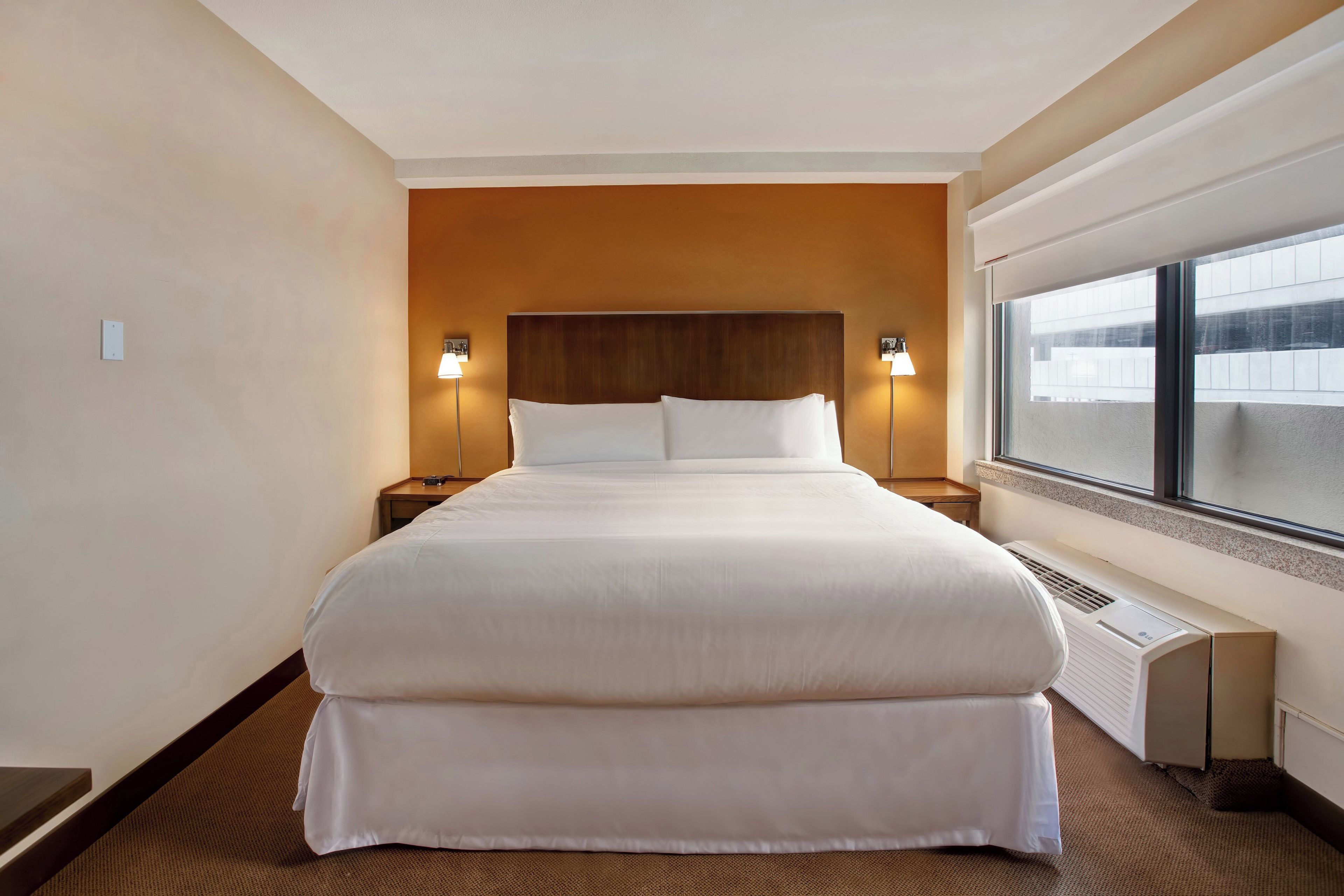 A hotel room with a large bed, two bedside lamps, a window, and a wall-mounted heater.