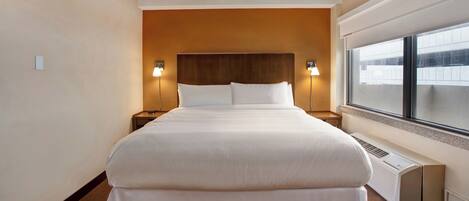 A hotel room with a large bed, two bedside lamps, a window, and a wall-mounted heater.