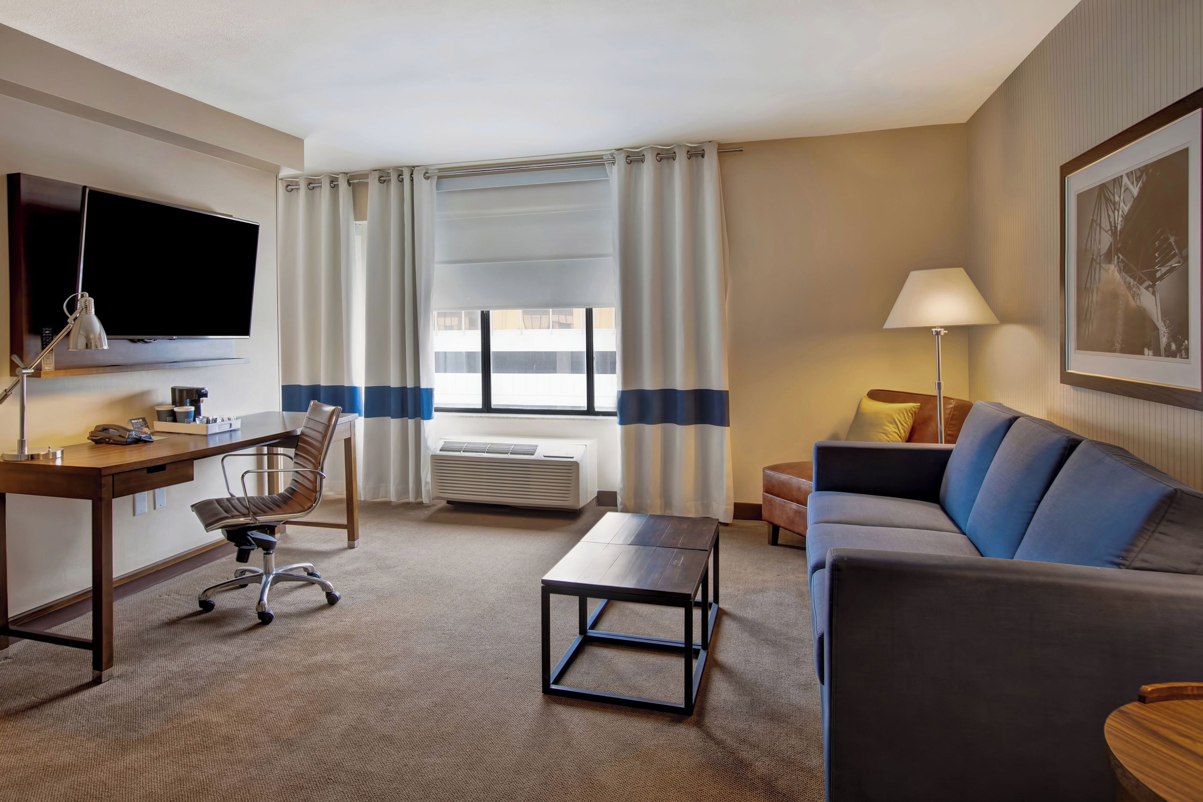 Une chambre d’hôtel équipée d’un bureau, d’une chaise, d’un canapé, d’une télévision, d’une lampe, d’un climatiseur et d’une fenêtre avec des rideaux.