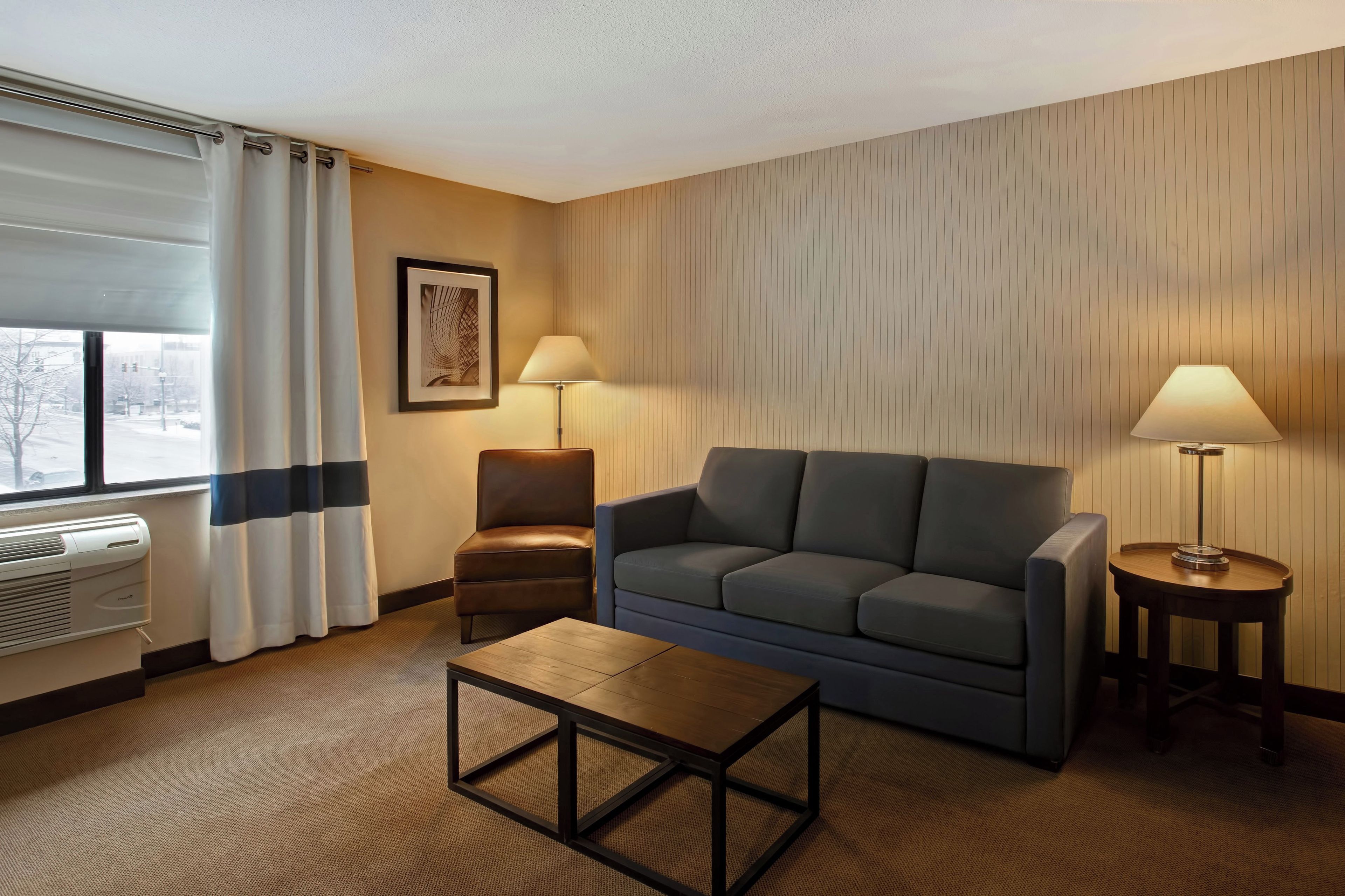 Habitación de hotel con sofá, dos mesitas de noche con lámparas, una mesa de centro, una silla, una ventana con cortinas y un cuadro colgado en la pared.