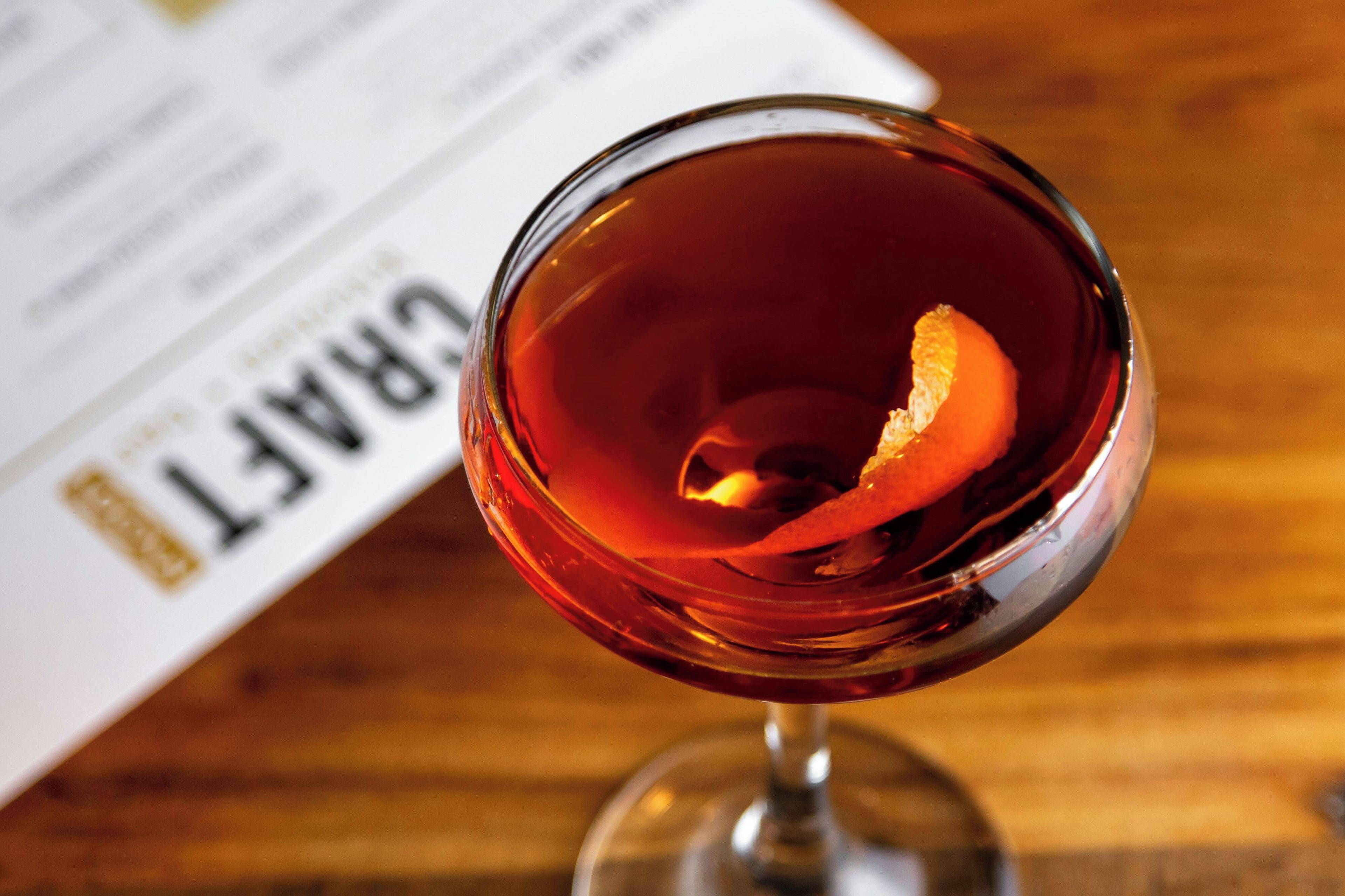 A glass with a dark amber liquid, possibly a cocktail, with a slice of orange peel on the rim.