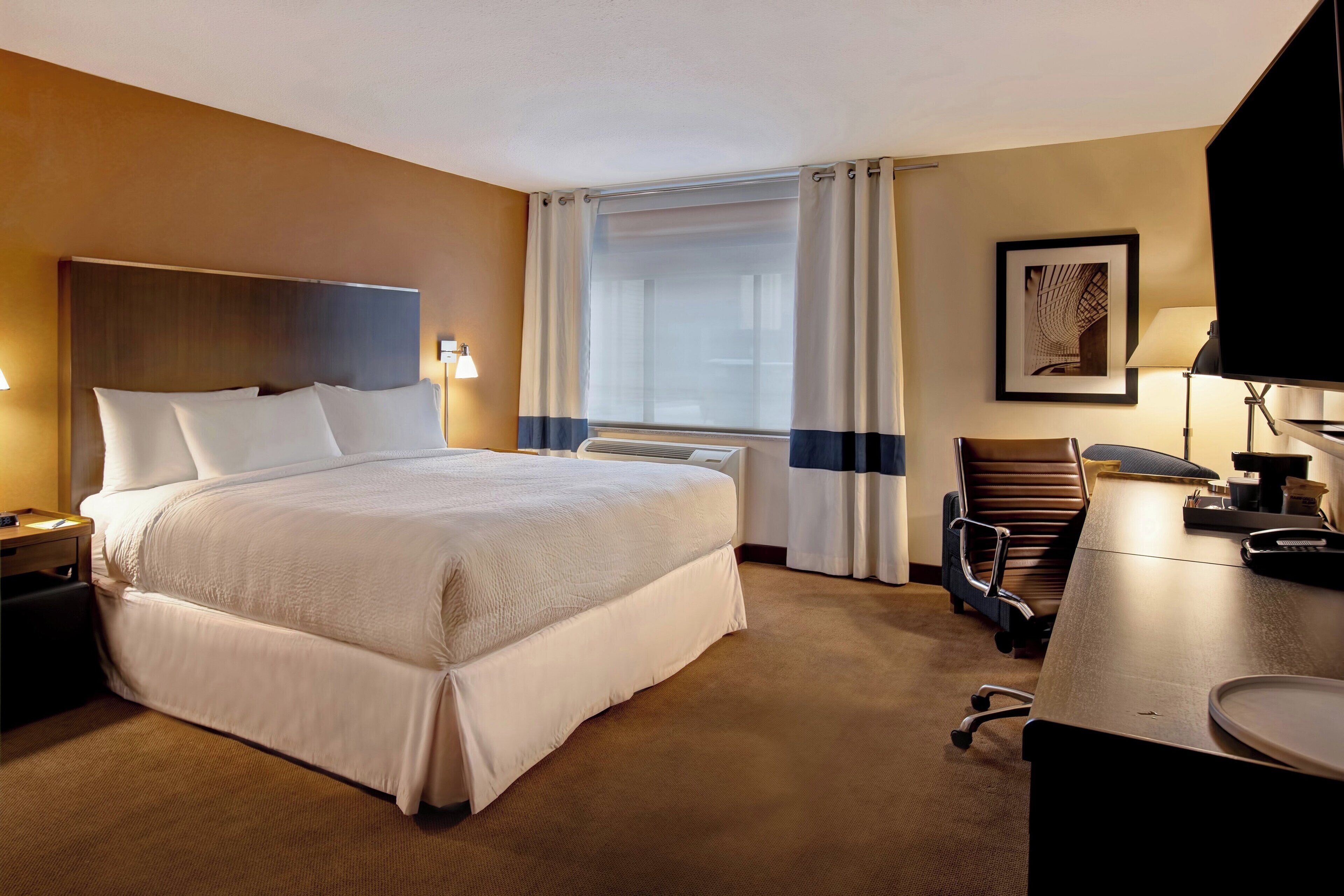 Une chambre d’hôtel avec un grand lit, un bureau, une chaise, une lampe de bureau et une fenêtre avec des rideaux.