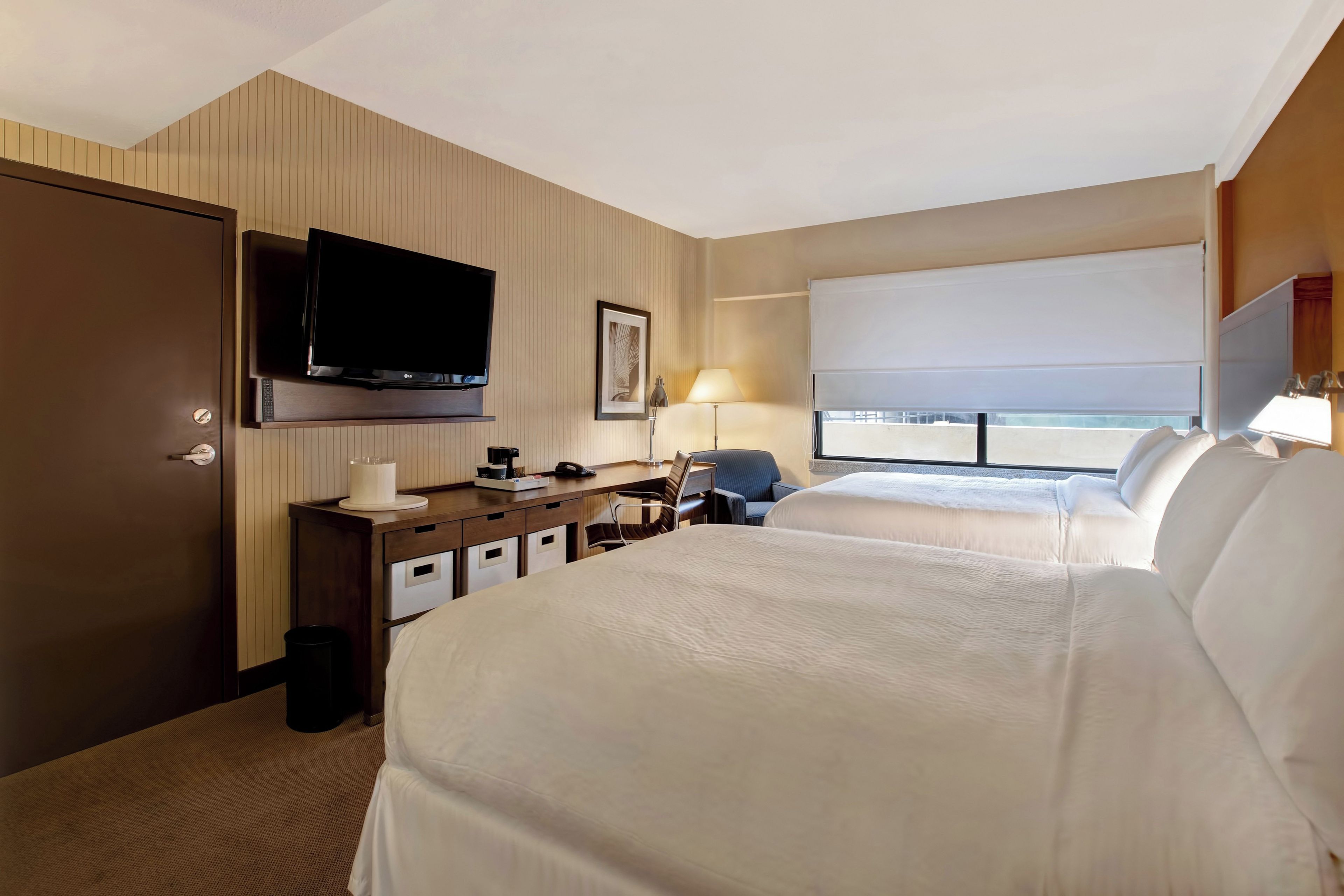 A hotel room with a bed, a desk with a television, a chair, a window with a view, and a door.