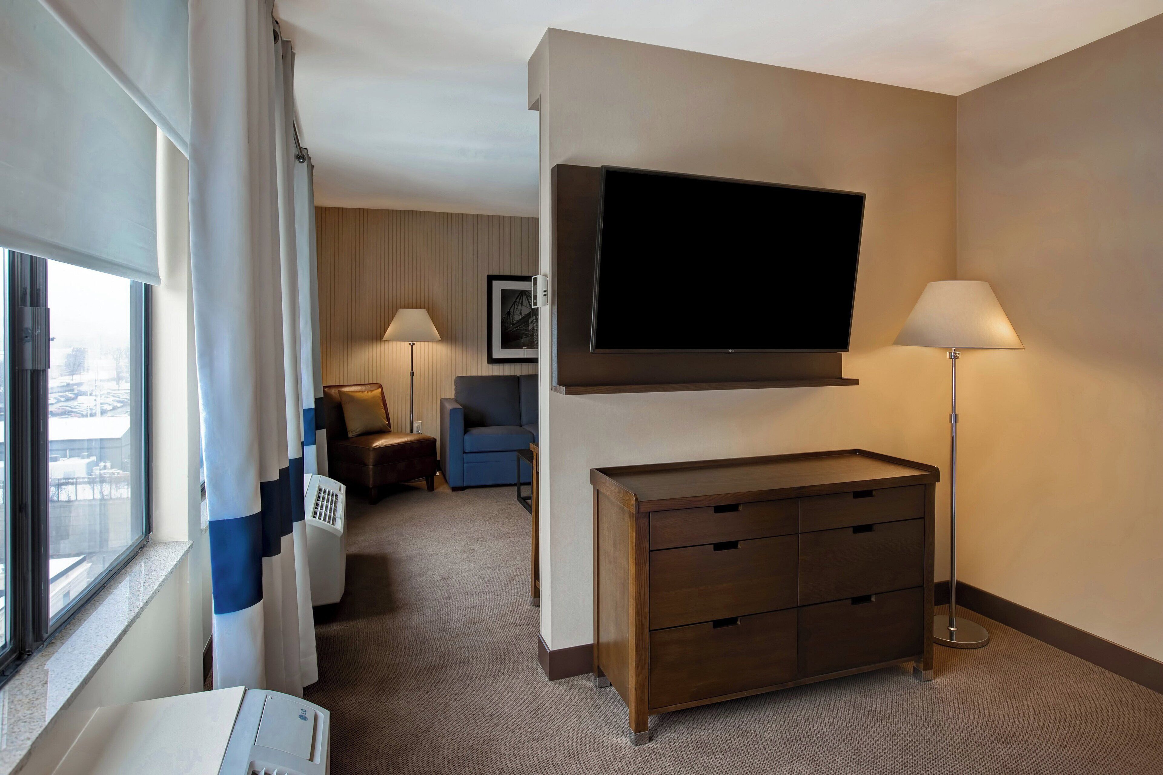 A hotel room with a large flat-screen TV mounted on the wall, a wooden dresser, a desk with a lamp, a sofa, and a window with curtains.