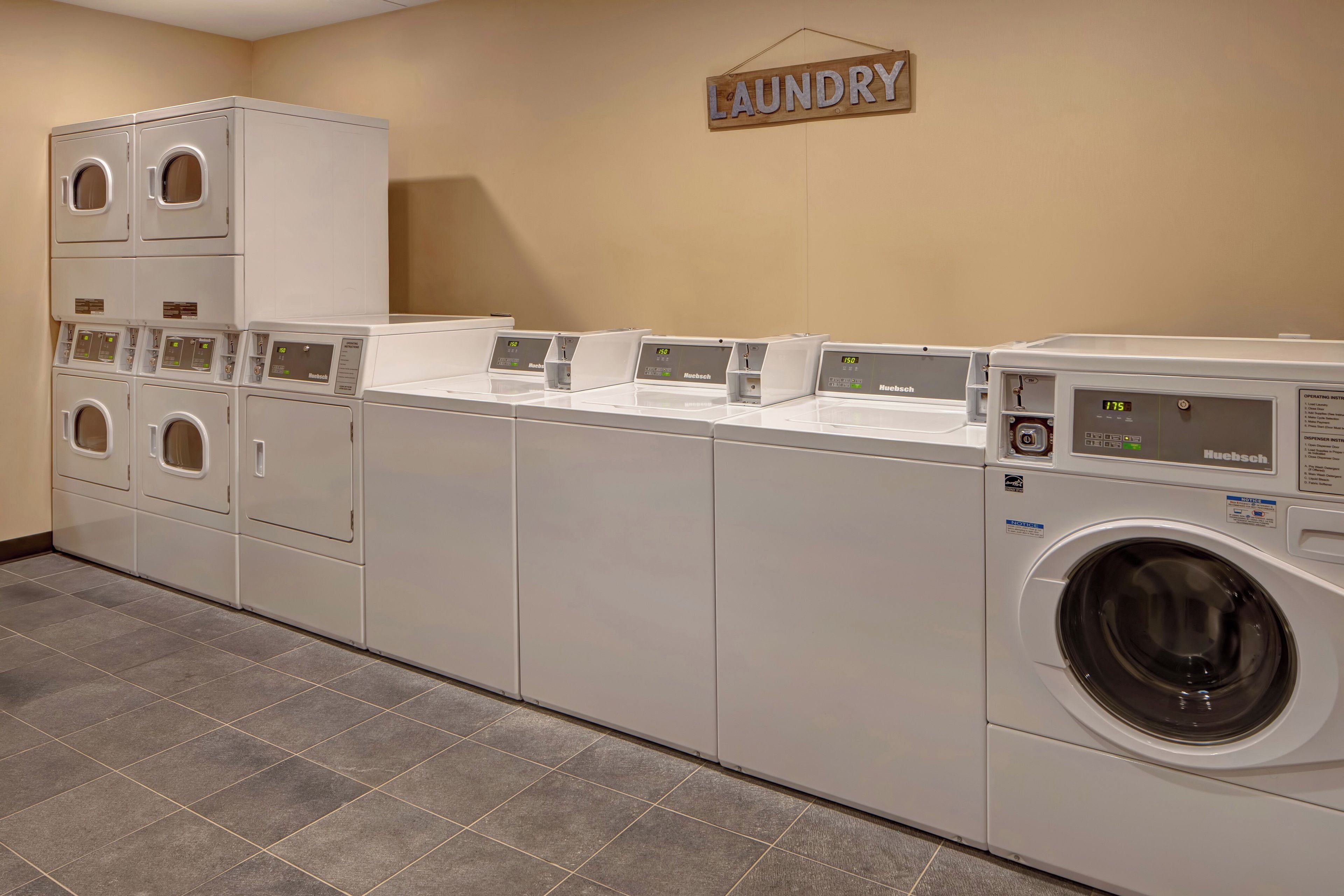 A laundry facility with multiple washing machines and dryers, a wall-mounted sign indicating the area, and a tiled floor.