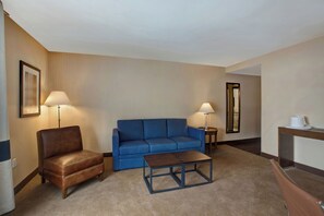 A hotel room with a brown leather chair, a blue sofa, a wooden coffee table, two floor lamps, and a mirror on the wall. - Four Points By Sheraton Peoria (Peoria)