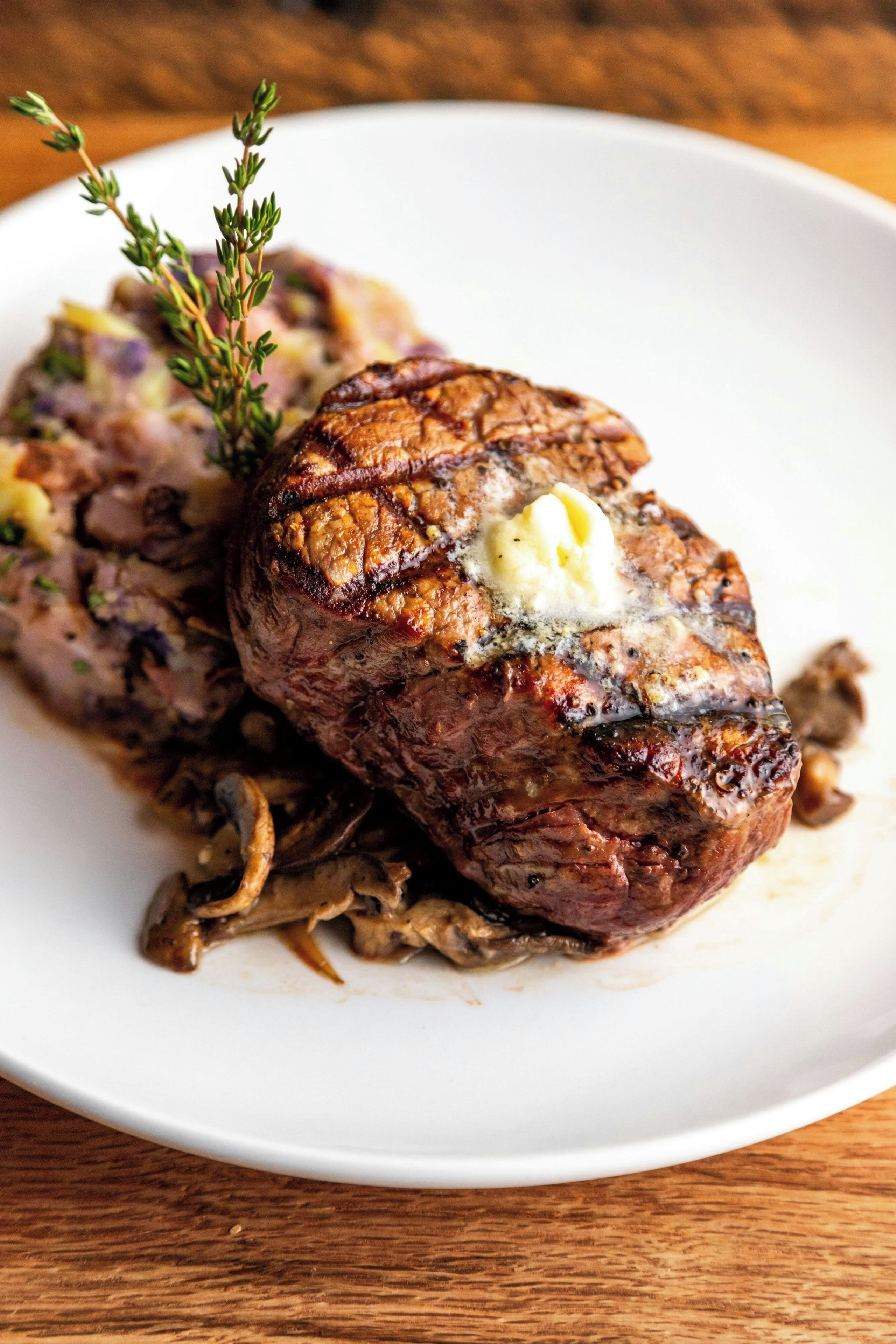 A steak with a pat of butter on top, served on a white plate with mushrooms and a sprig of thyme.